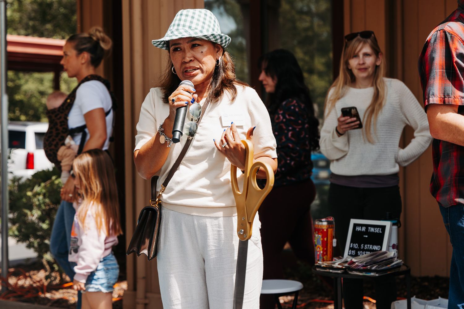 Woman in white outfit and bucket hat speaks into microphone at an outdoor community event.