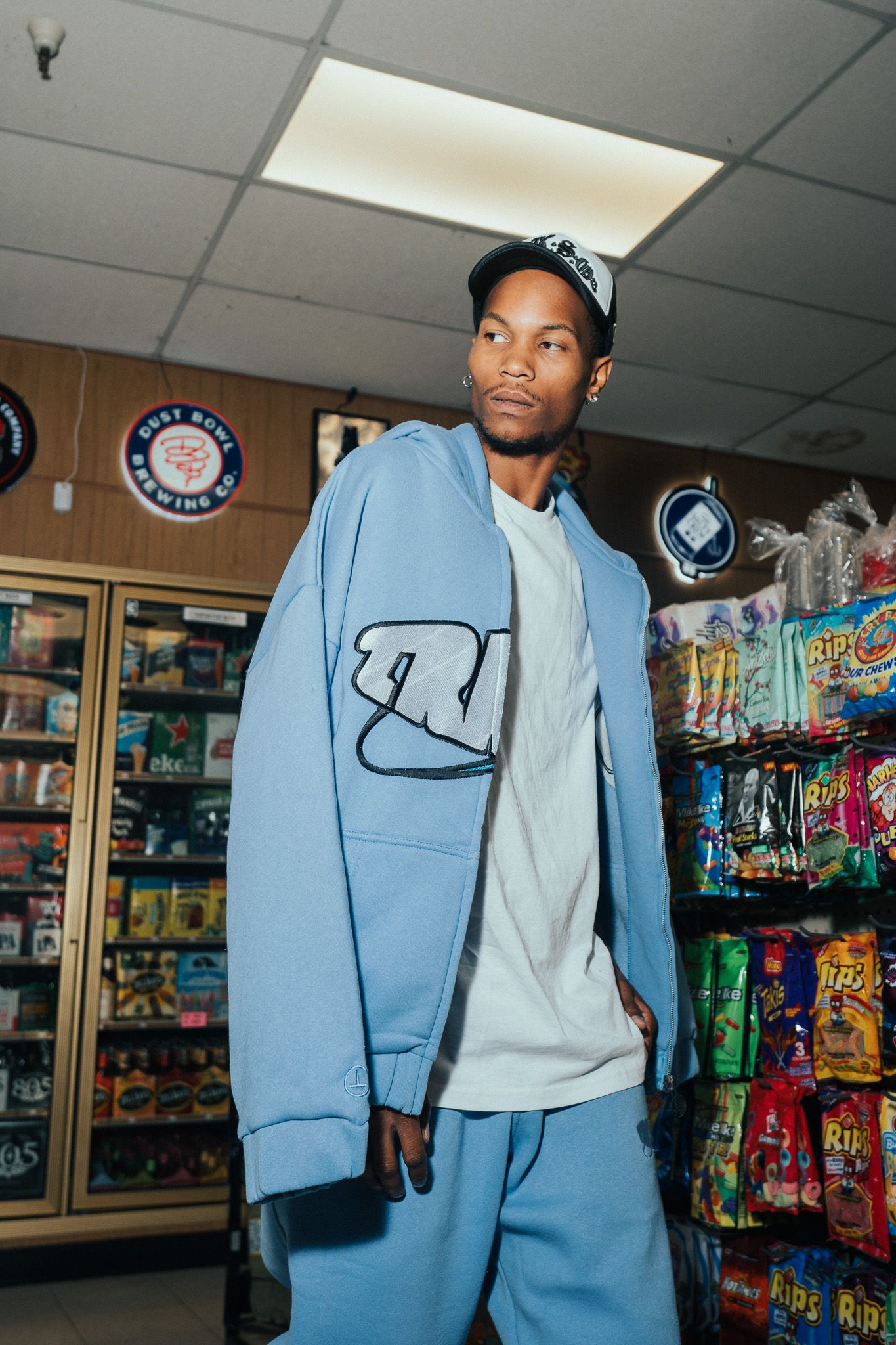 Young man in blue zip-up hoodie and matching pants poses in a convenience store snack aisle.