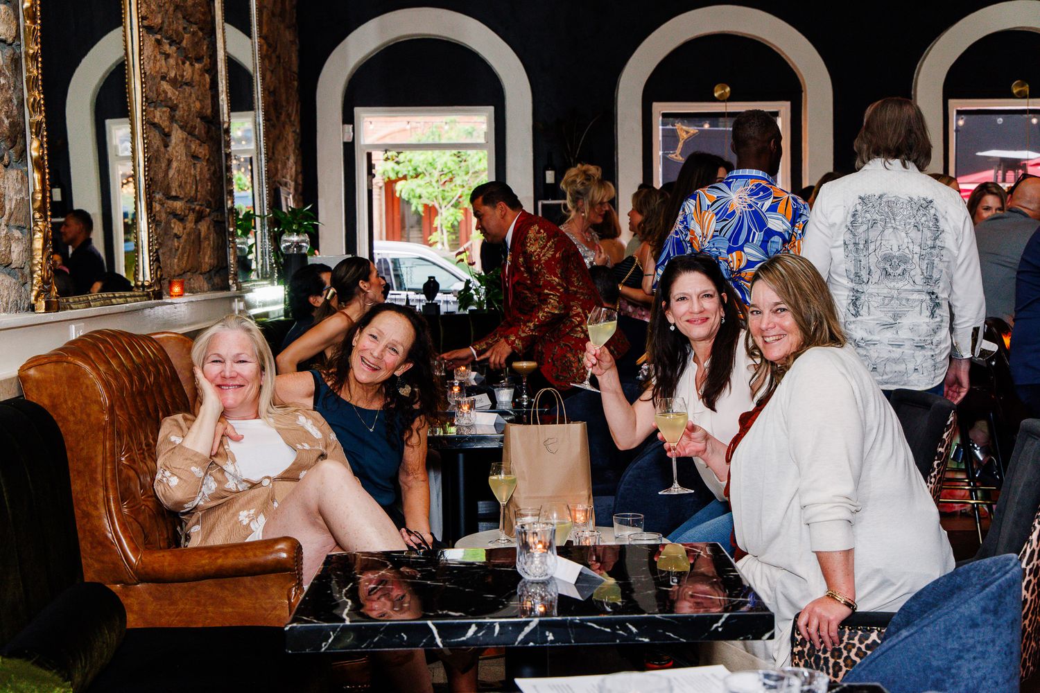 Four women smiling and toasting with wine glasses at an elegant bar or restaurant social gathering.