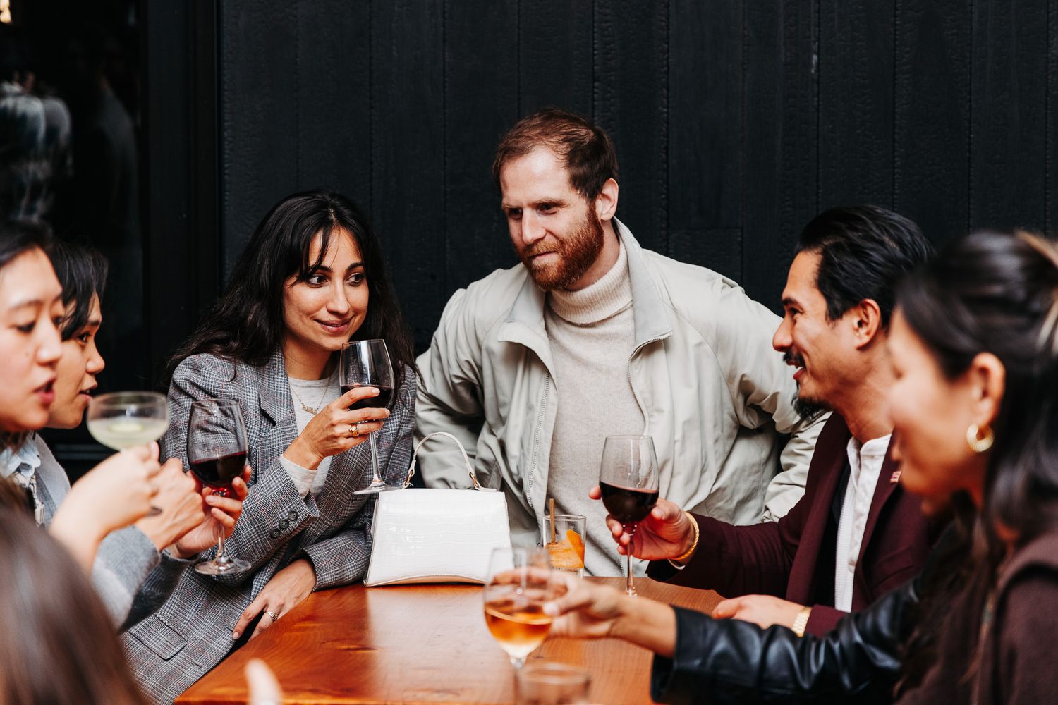 A group of friends gather around a table sharing drinks and conversation in a dimly lit setting.