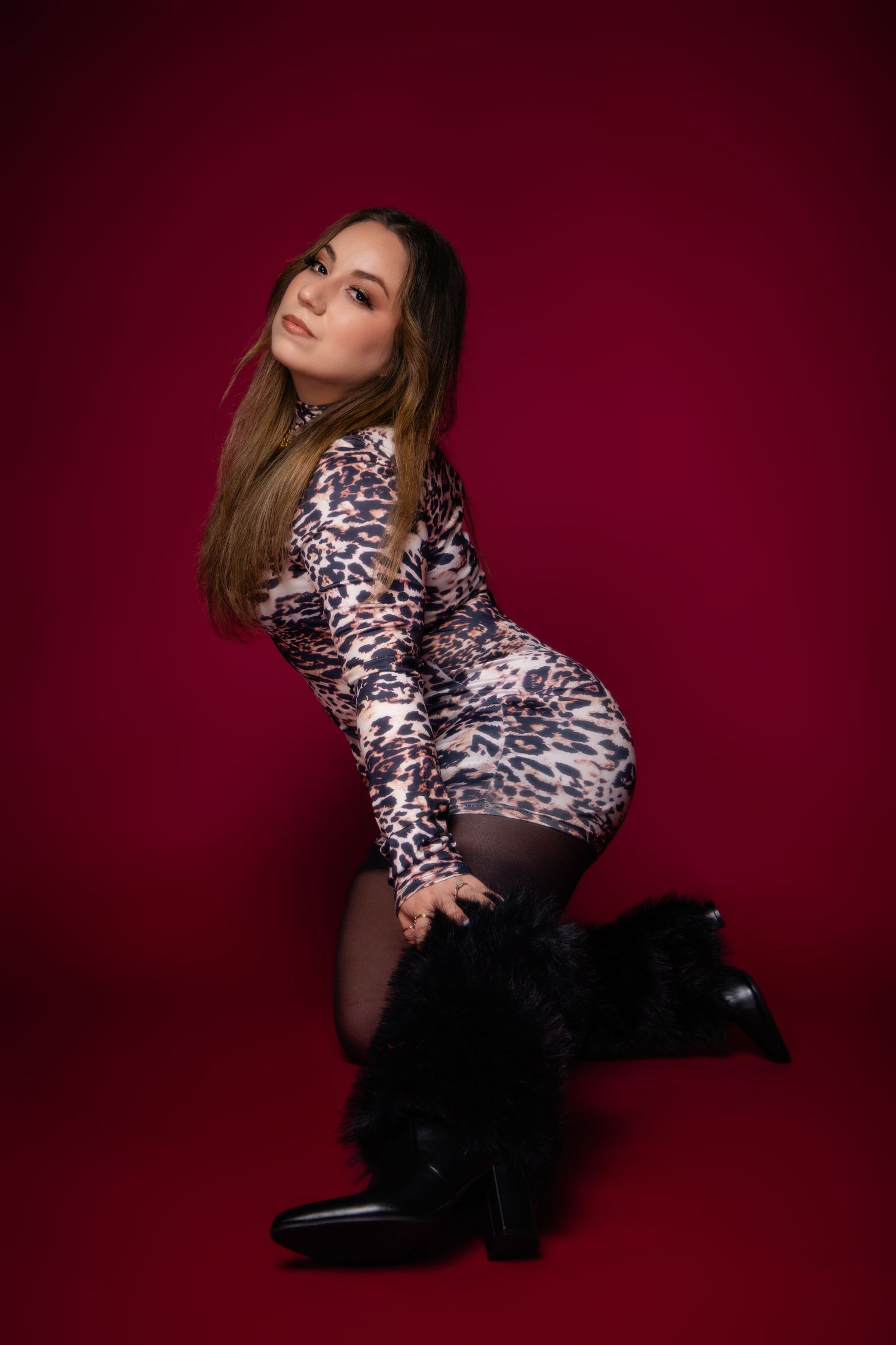 Model in leopard print dress and black fur boots poses against burgundy background in studio setting.