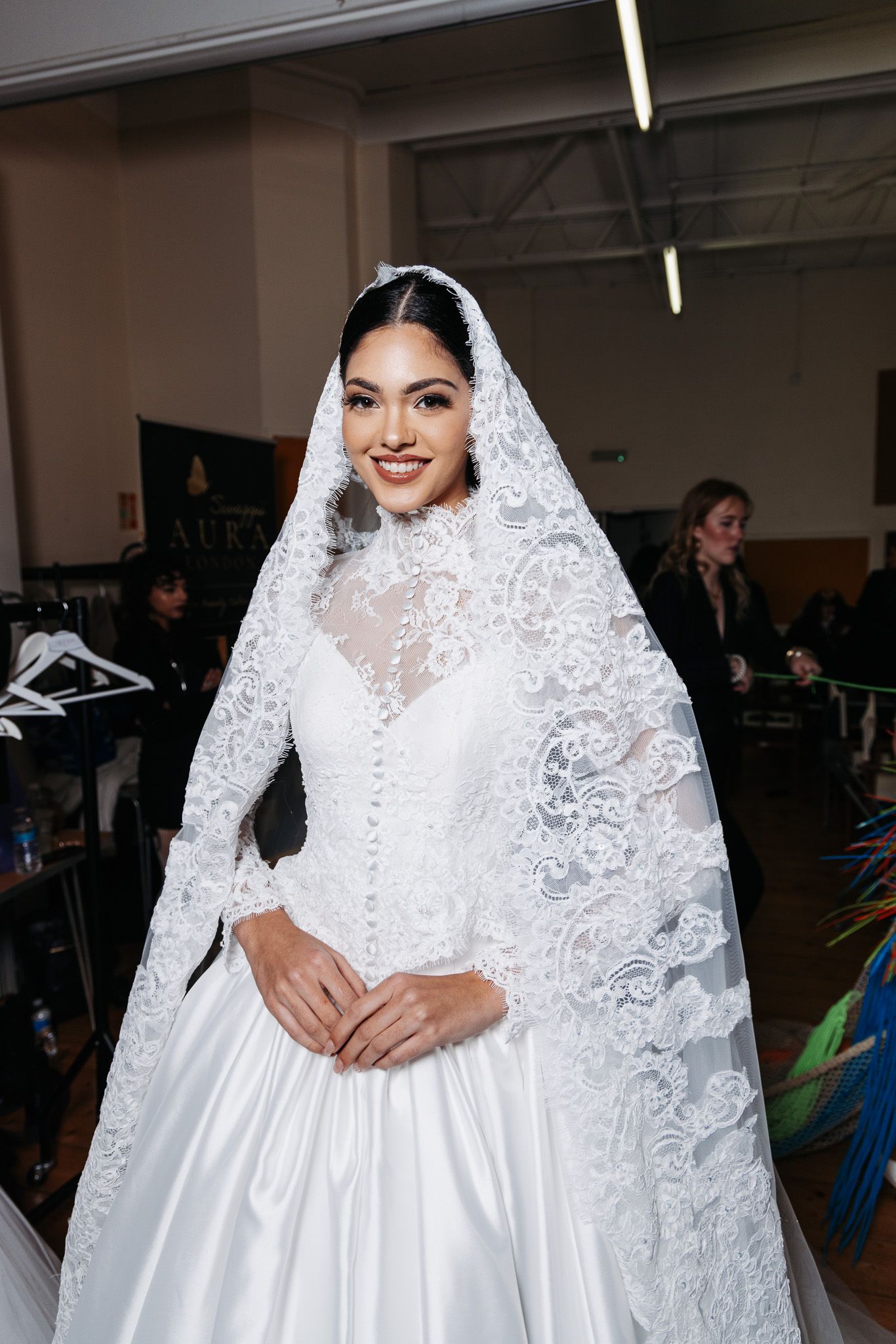 A bride in a stunning white lace wedding dress with a long veil poses in an indoor setting.