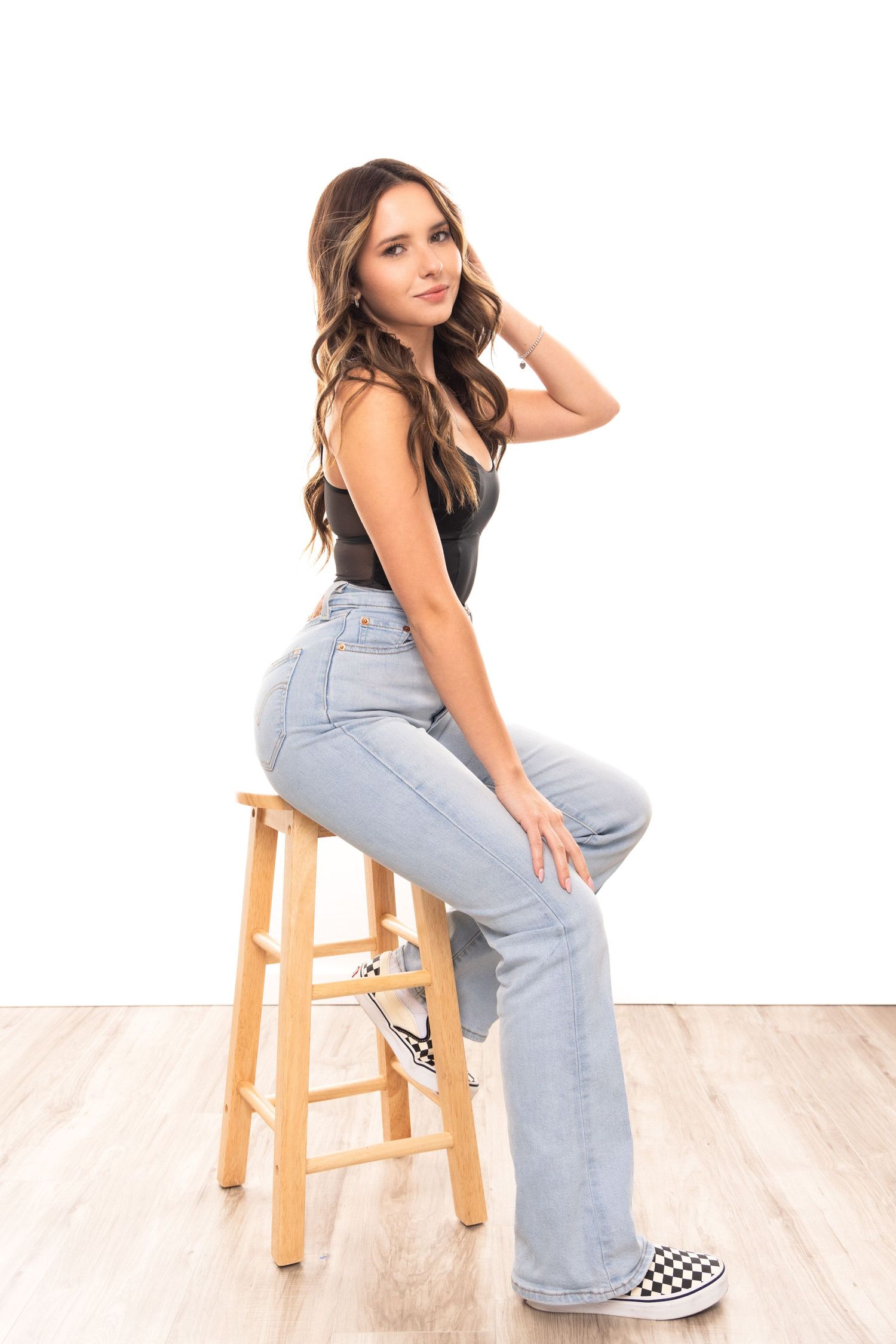 Person in light blue jeans and checkered Vans shoes poses casually on wooden stool against white wall.
