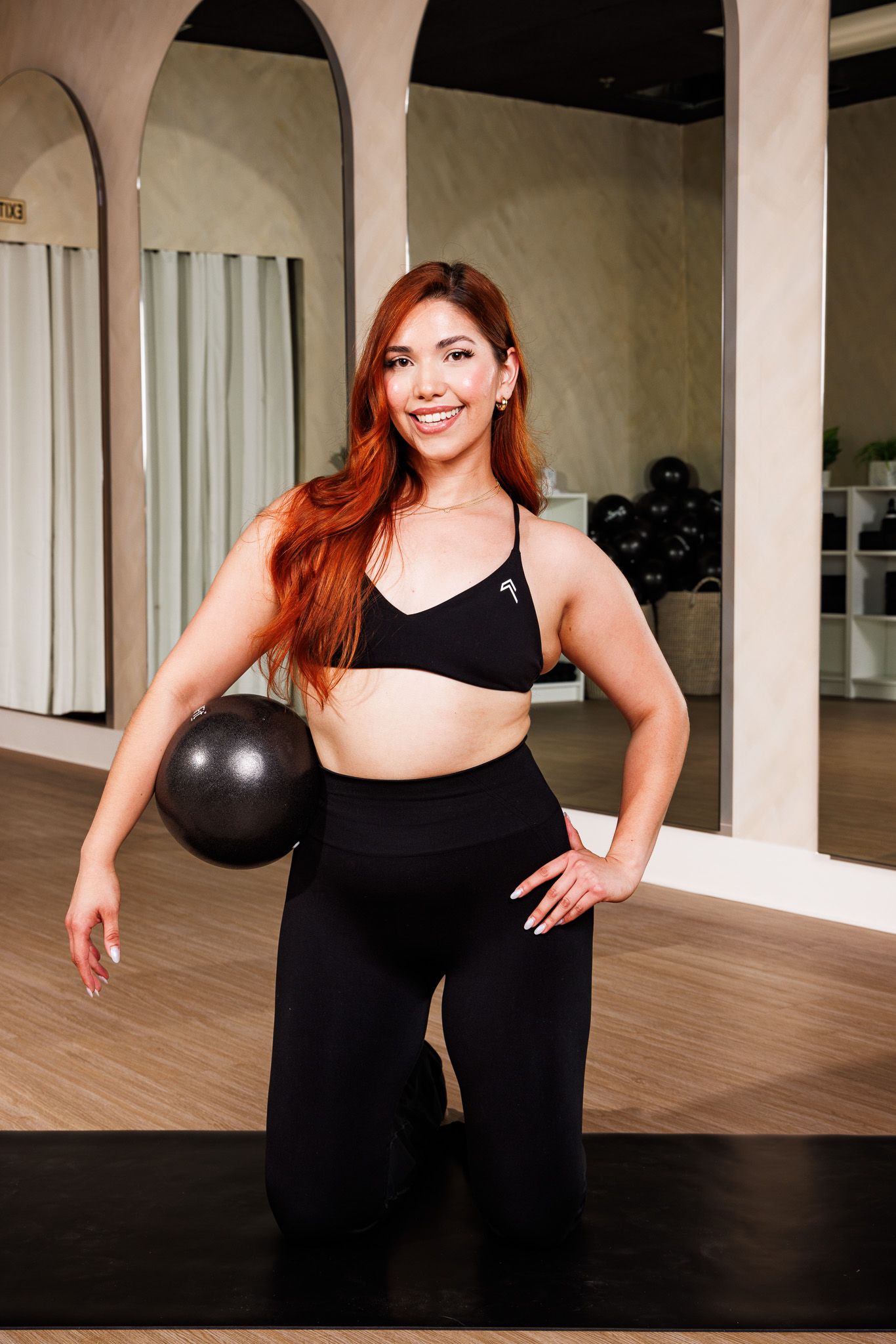 Smiling woman with red hair in black sports bra and leggings holding a small ball in a gym studio.