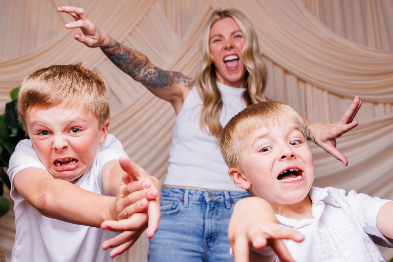 Woman with tattoos poses playfully with two young blonde boys making funny faces and reaching toward camera.