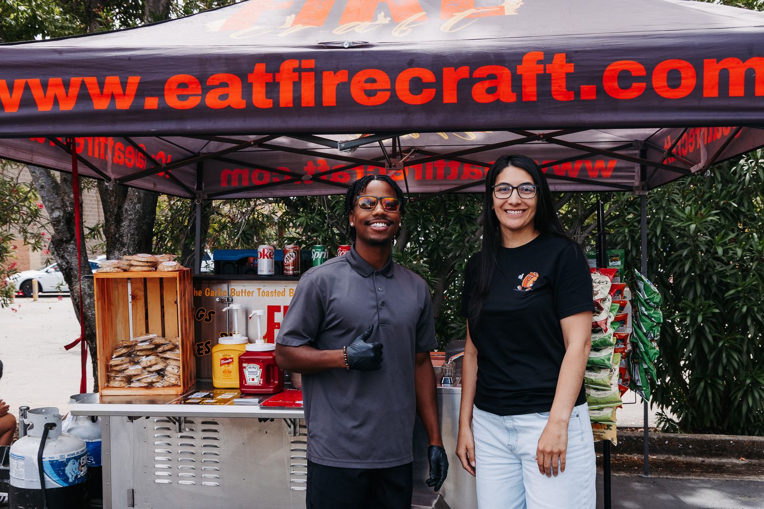 Two vendors smile at eatfirecraft.com food stand under orange canopy at outdoor market event.