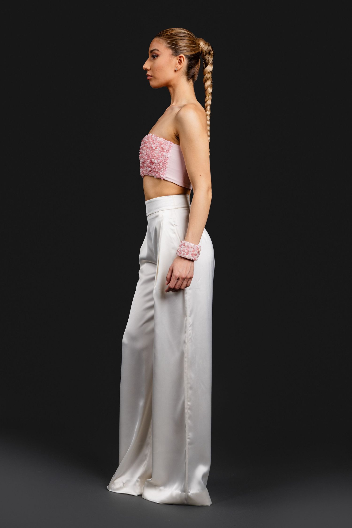 Model in a pink floral strapless crop top and white wide-leg satin trousers with matching pink bracelet, side view.