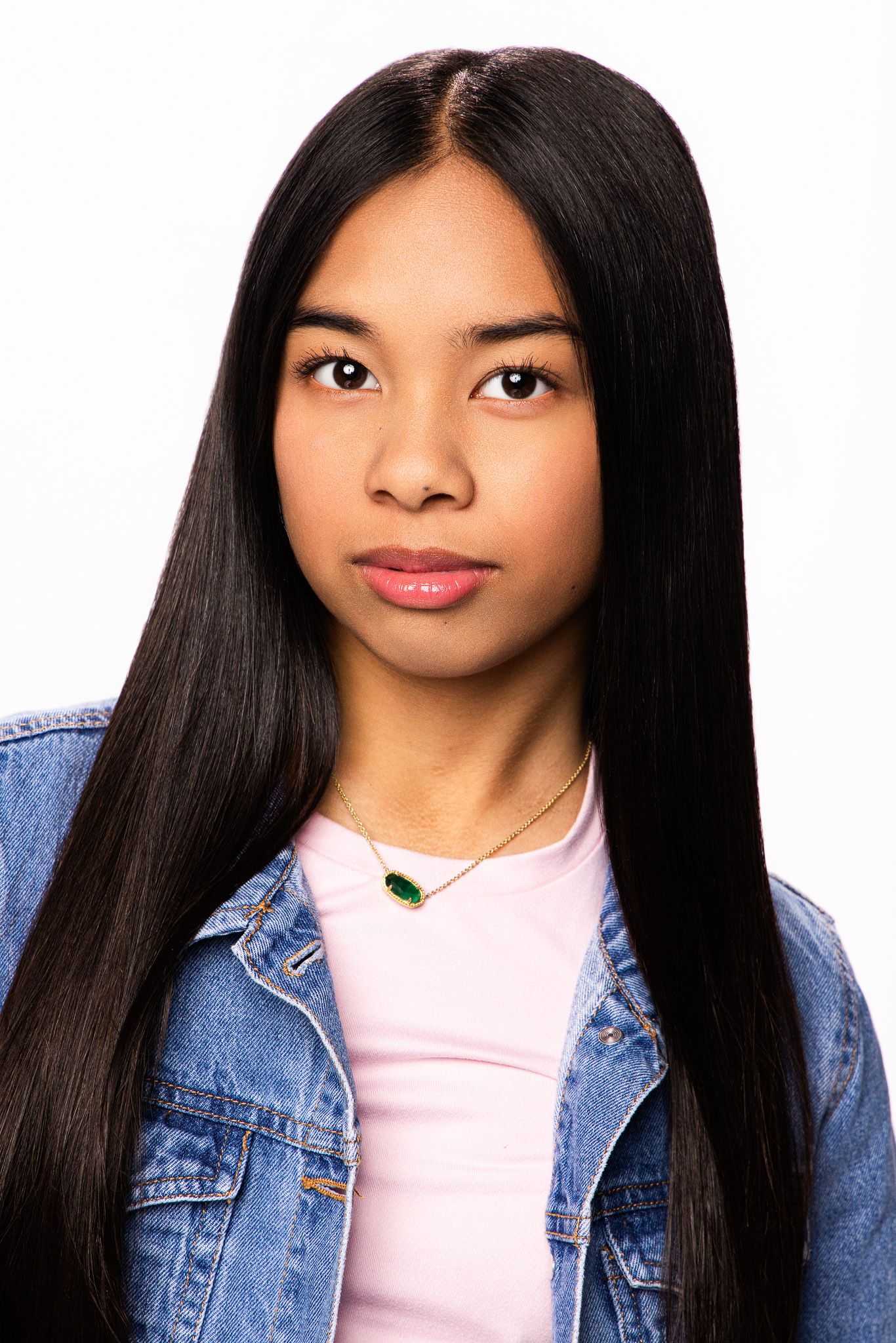Young woman with long straight black hair wearing a denim jacket and pink top with a green pendant necklace.
