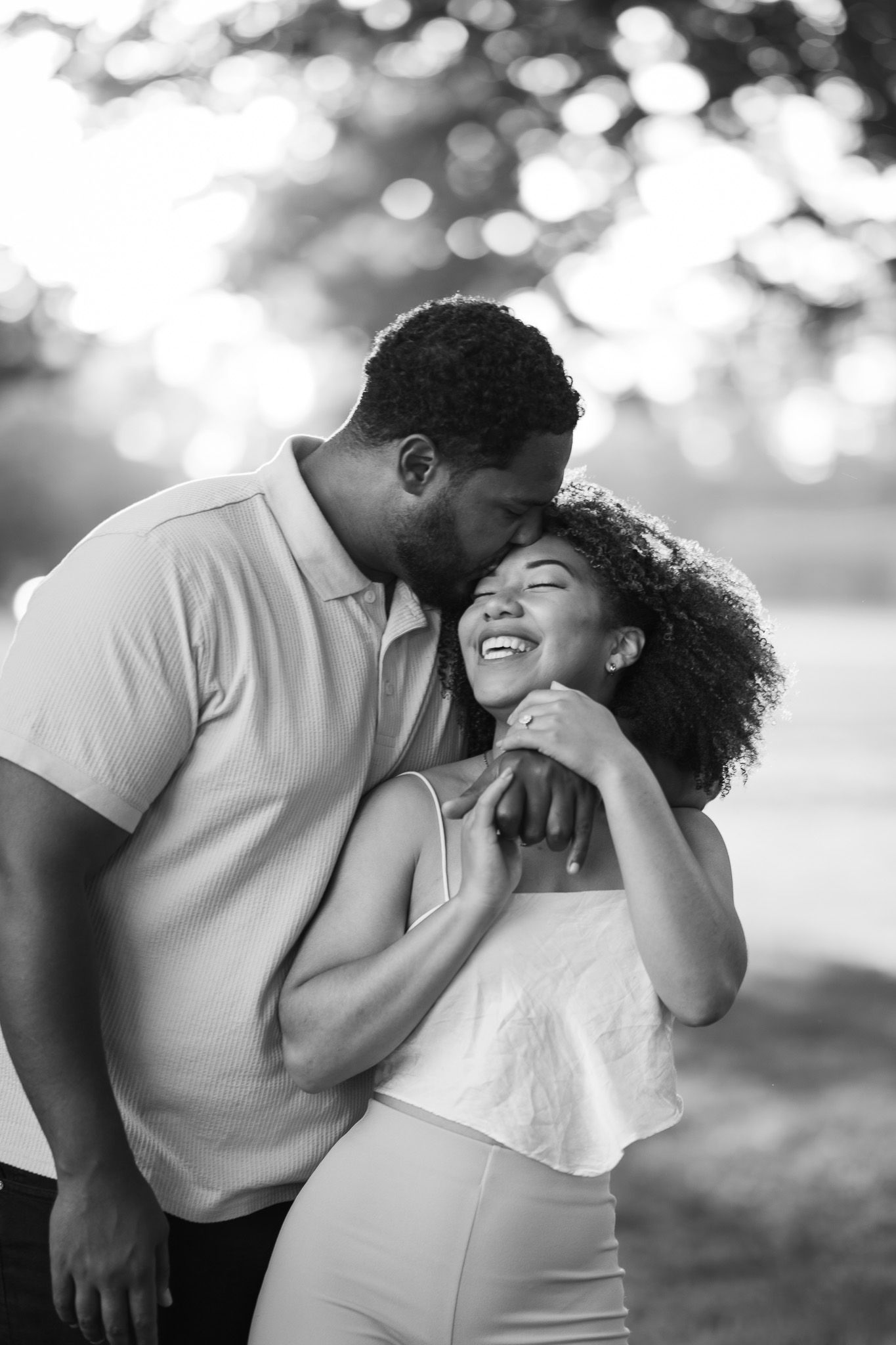 A tender black and white portrait of a couple sharing an intimate moment outdoors with bokeh lighting in the background.