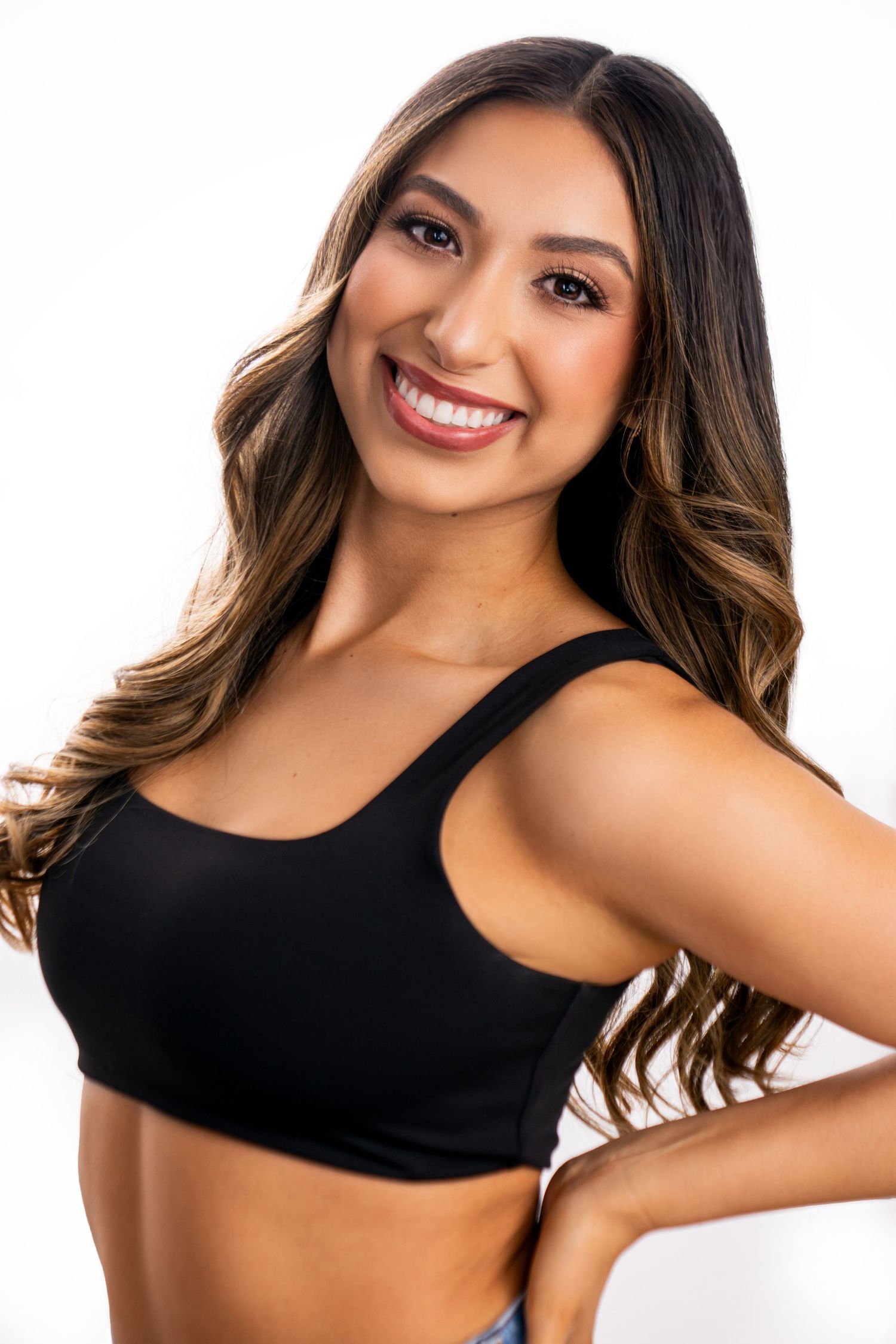 Young woman with wavy brown hair wearing black crop top smiling at camera against white background.