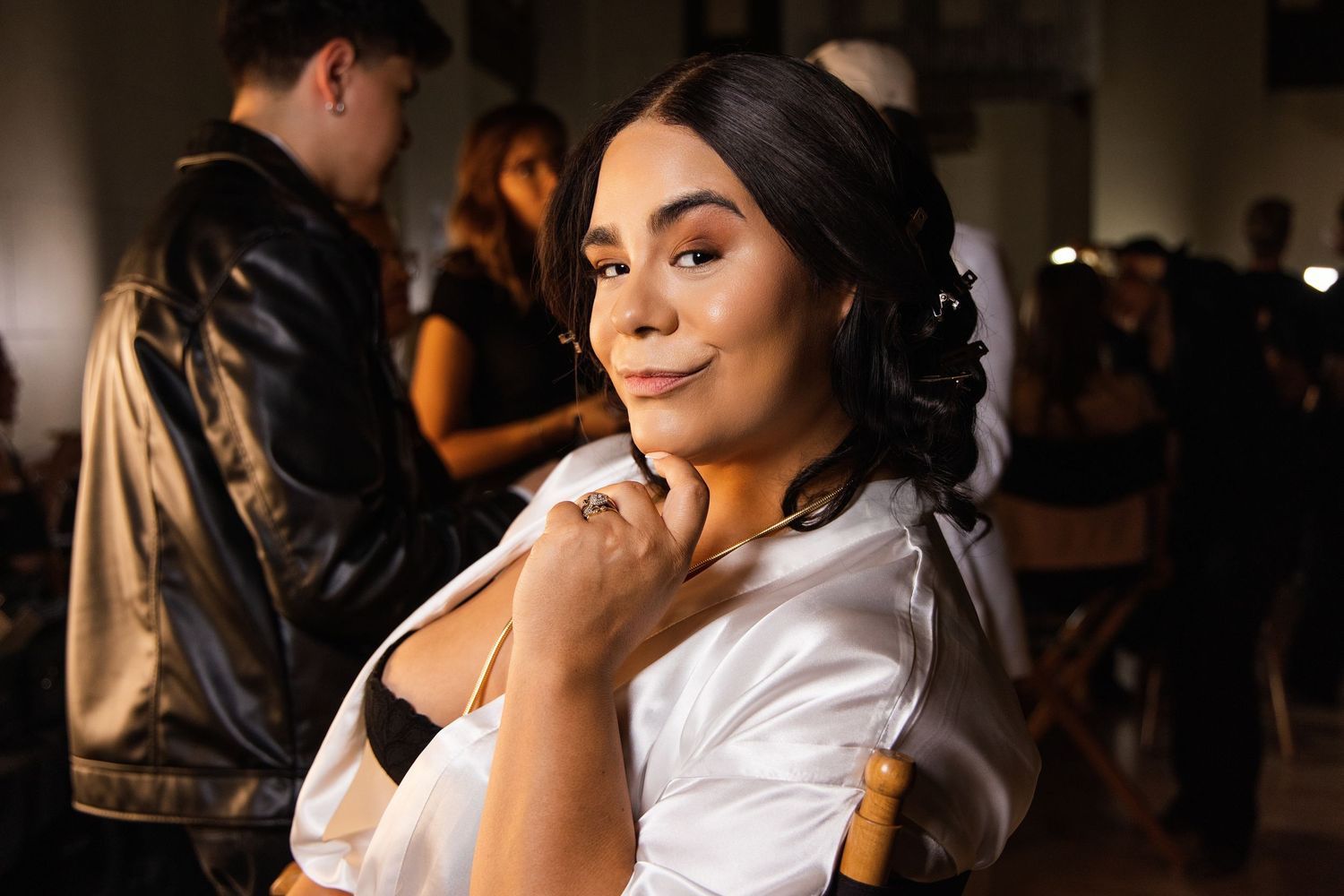 A woman in a white satin robe poses backstage, smiling at the camera with elegant updo hairstyle.