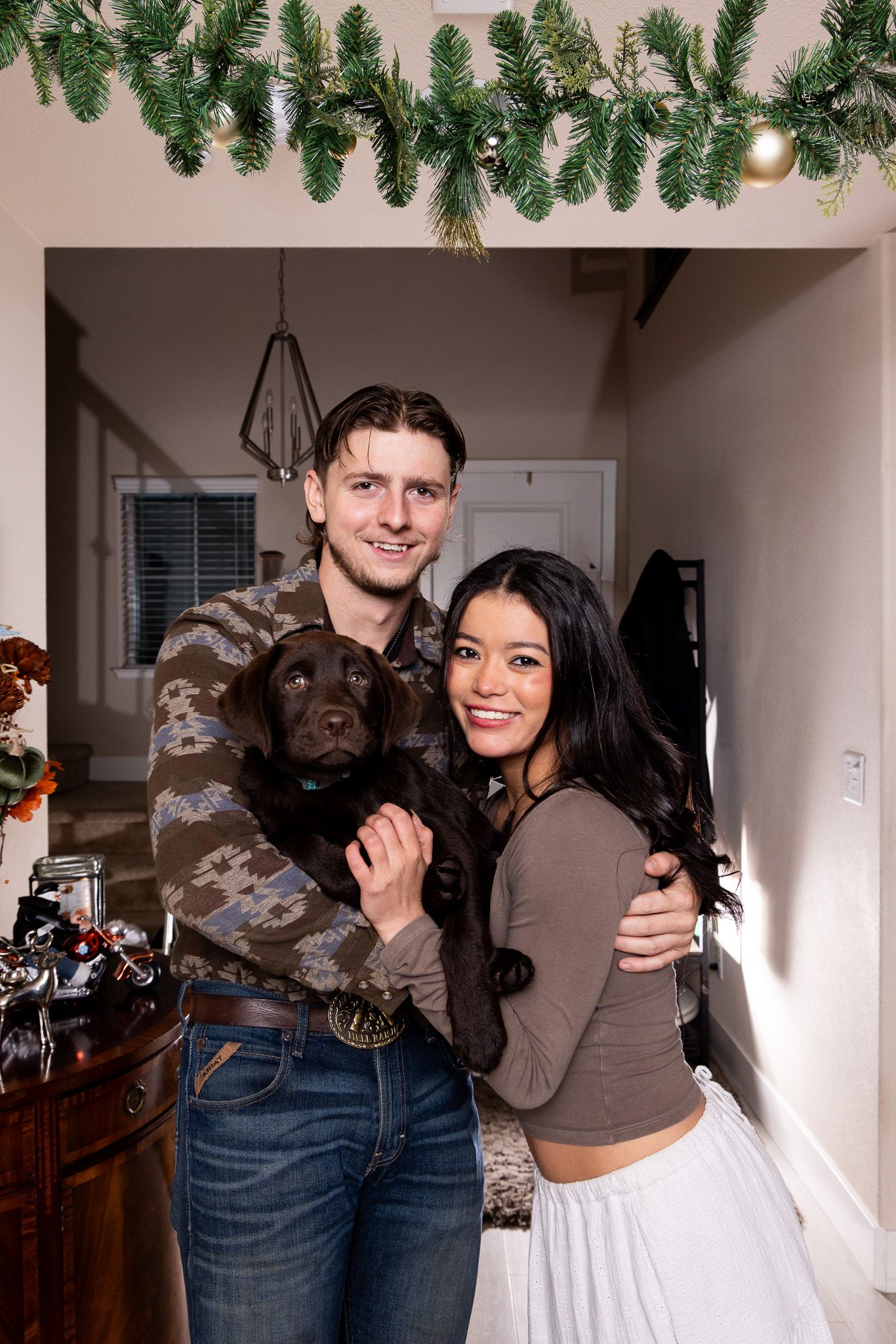 A couple poses together in front of Christmas decorations in a cozy indoor setting.
