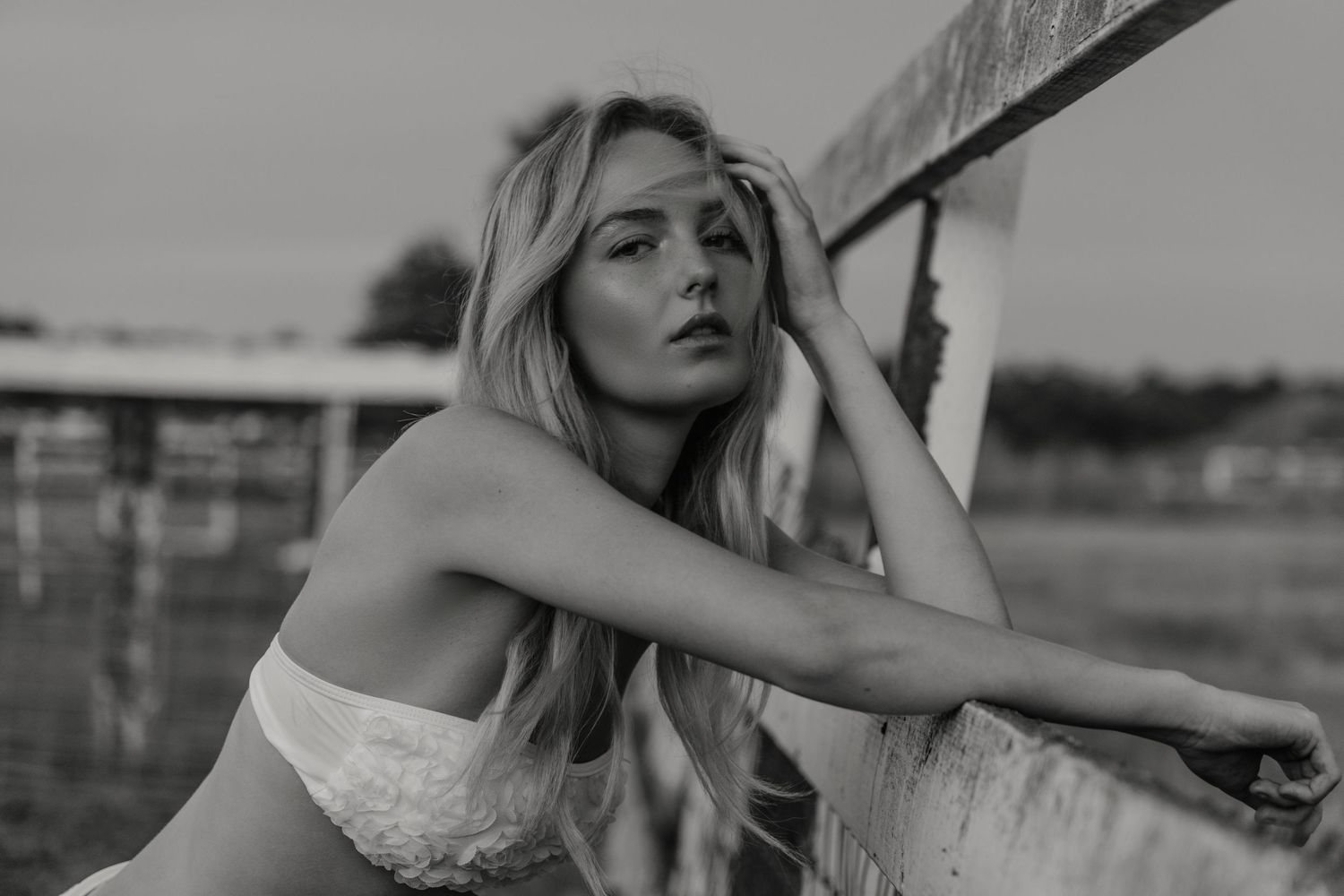 Black and white photo of a blonde woman leaning on a railing outdoors, gazing pensively into the distance.