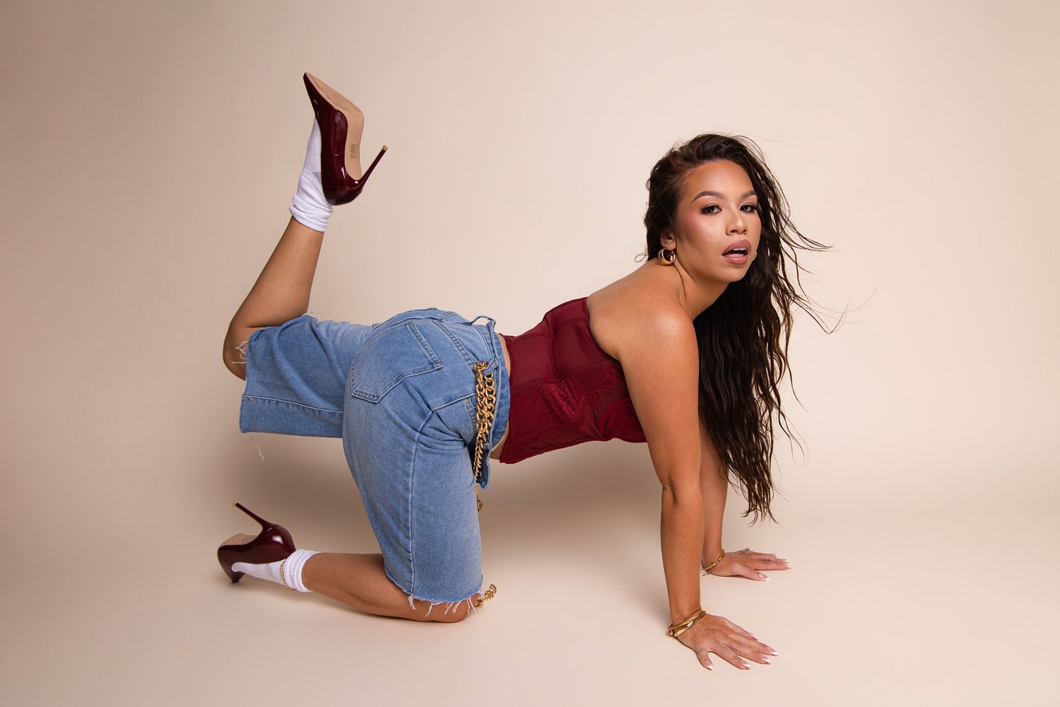 Woman in red top and jeans poses dynamically on studio floor wearing burgundy heels.