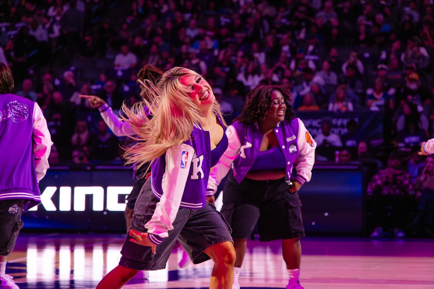 Sacramento Kings dancers performing energetically on court in purple varsity jackets during an NBA game.
