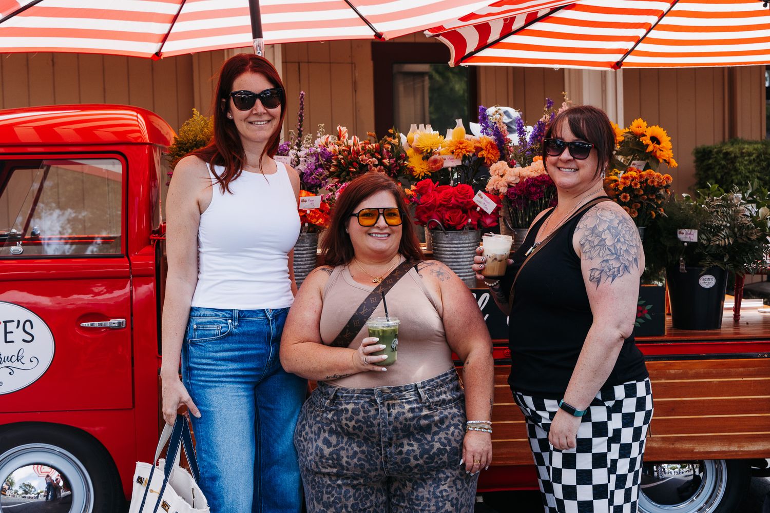 Three women posing together at an outdoor flower market beside a vintage red truck with colorful blooms.