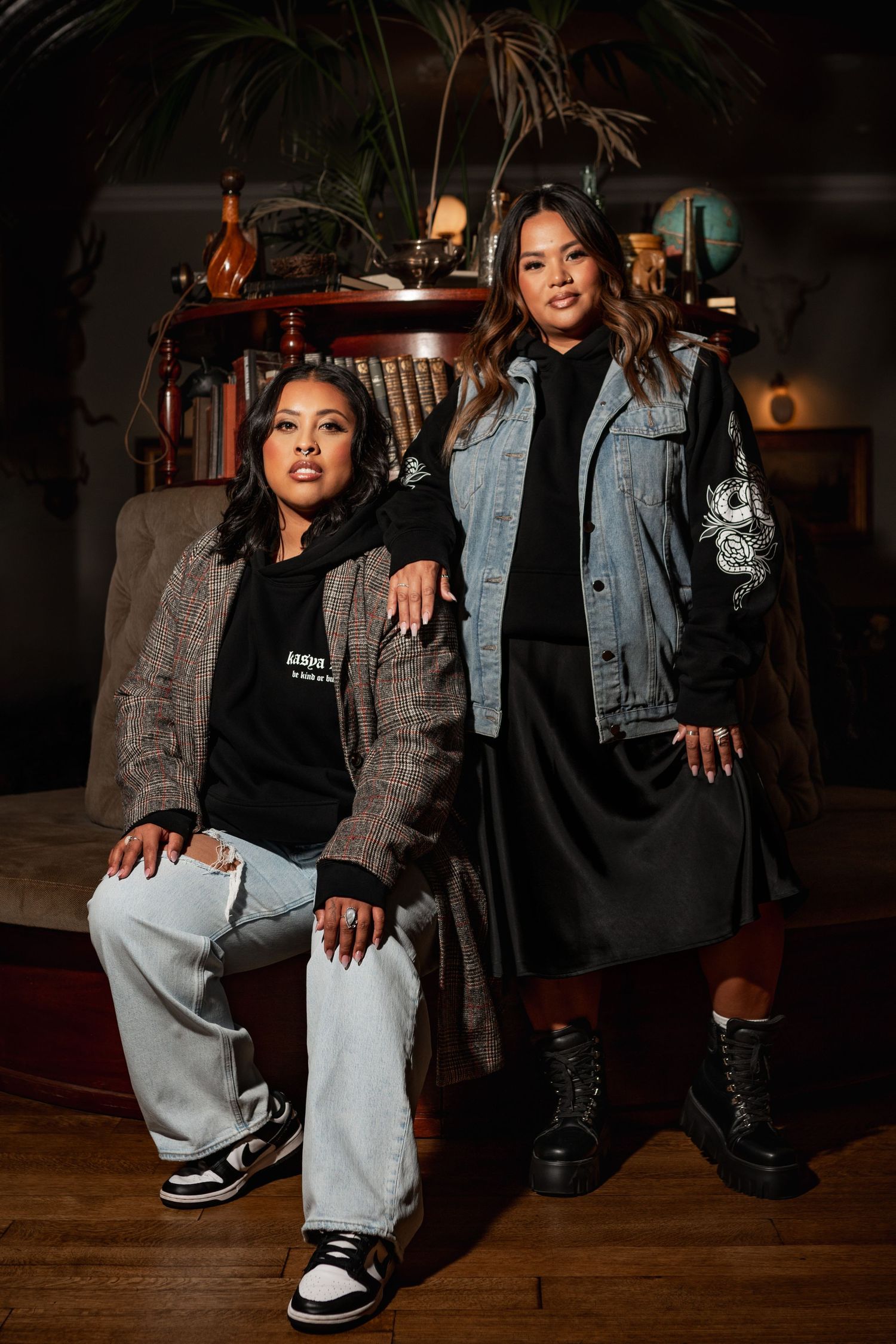 Two stylish women pose indoors, one seated in plaid blazer and Jordan sneakers, other standing in denim jacket and black skirt.