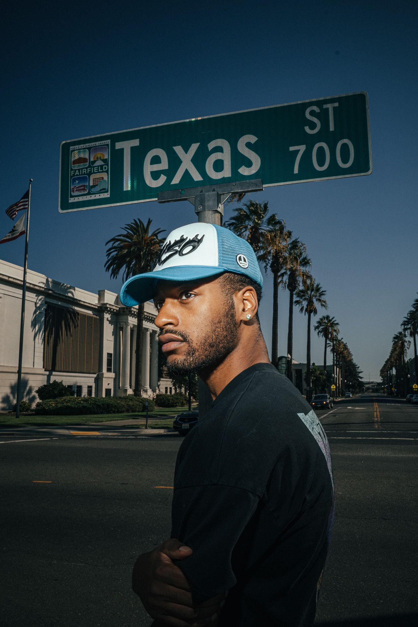 Man in blue cap poses under Texas St 700 sign with palm trees and blue sky in background.