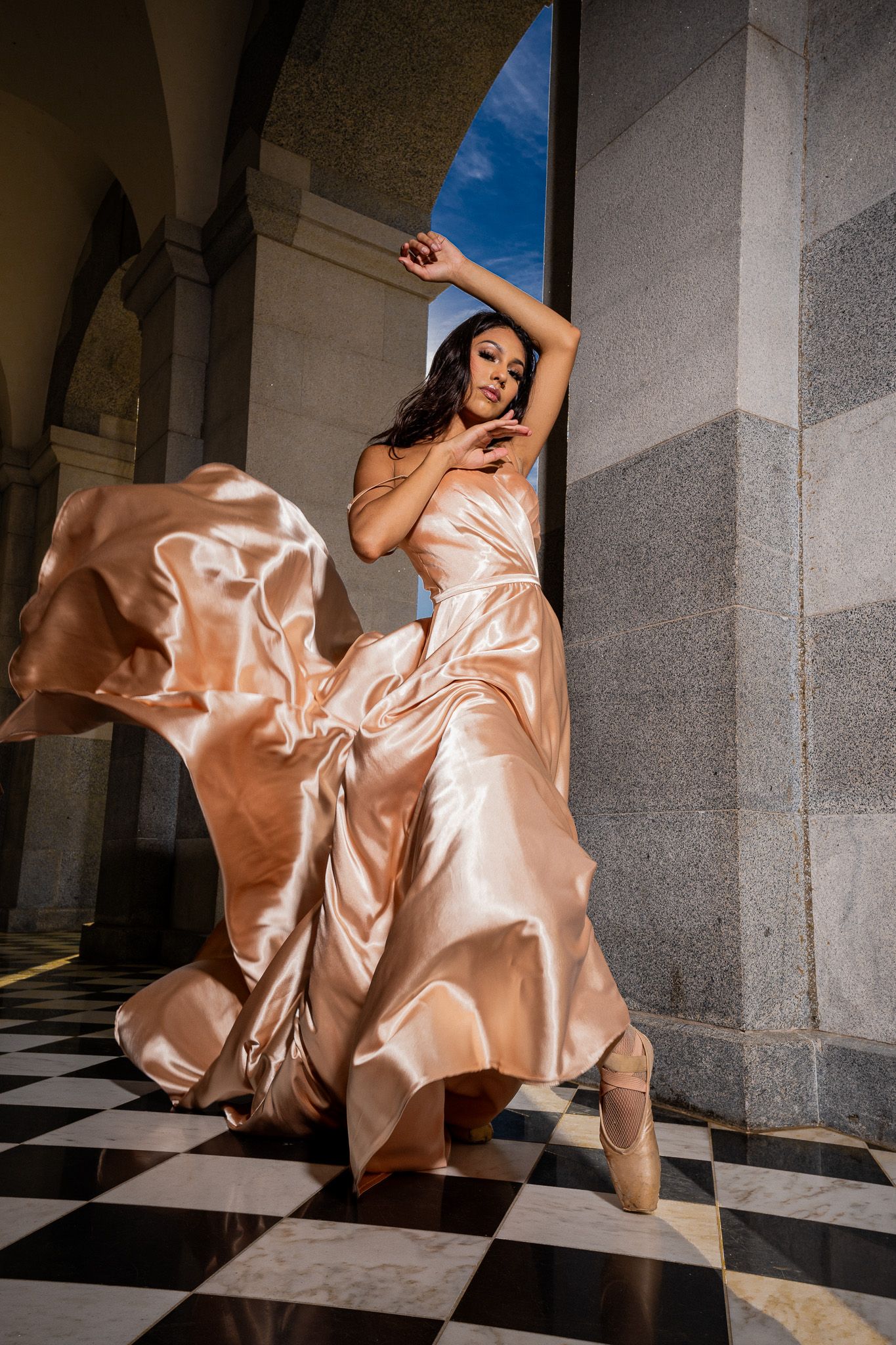 A dancer in a flowing champagne silk dress poses gracefully on a checkered floor beneath stone archways.