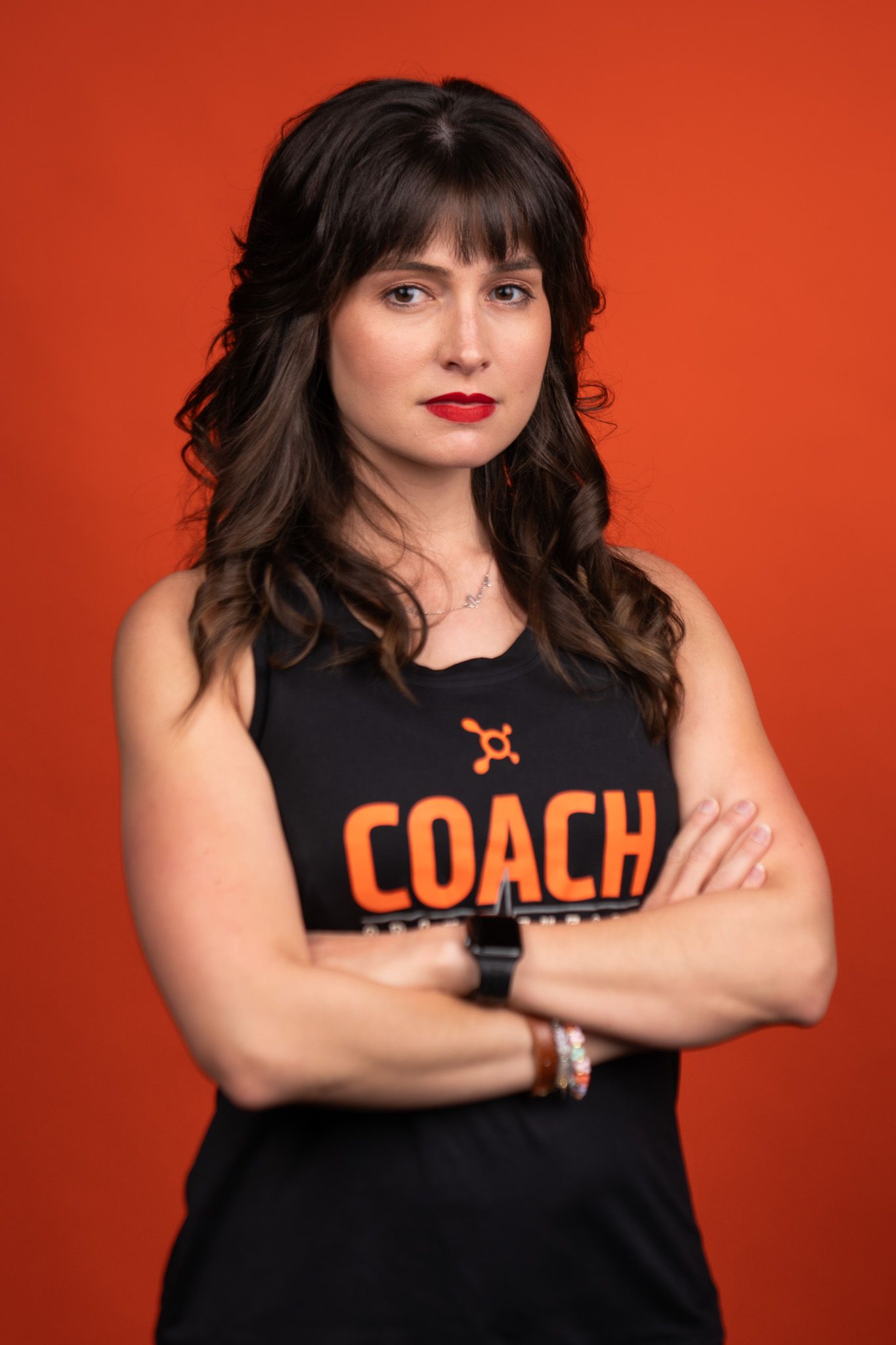 A person in a black COACH tank top poses with crossed arms against a vibrant orange background.