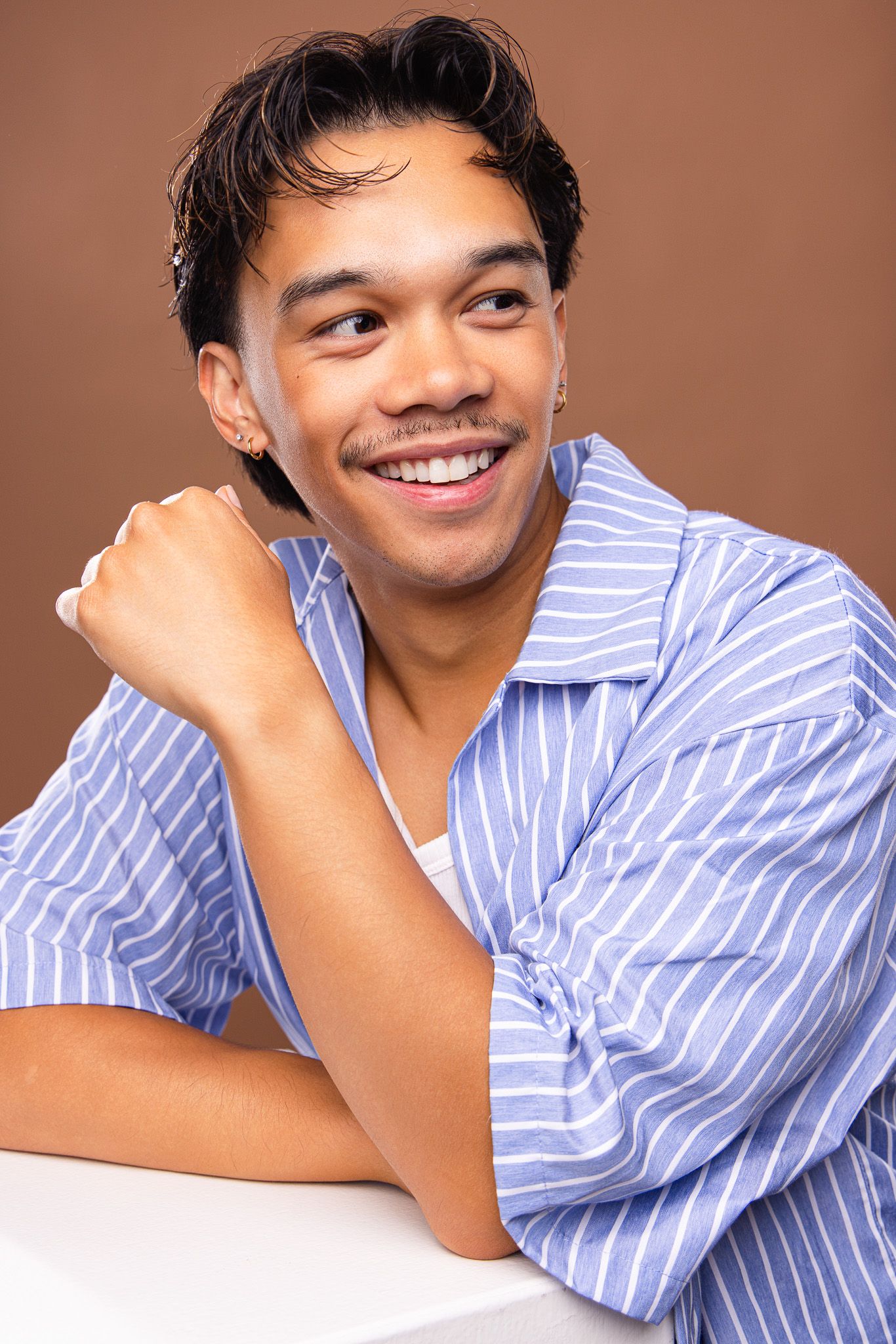 Young man in blue striped shirt smiling confidently against brown background, resting chin on hand.