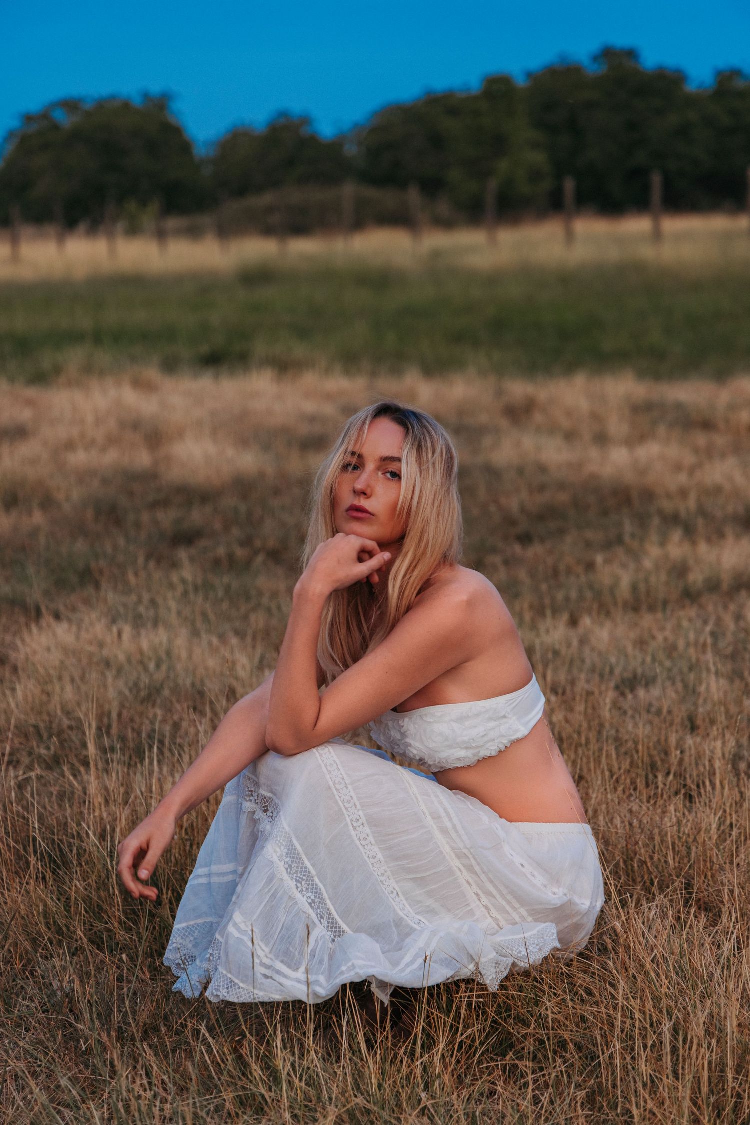Blonde woman in white crop top and skirt sitting in golden field with green trees and blue sky.