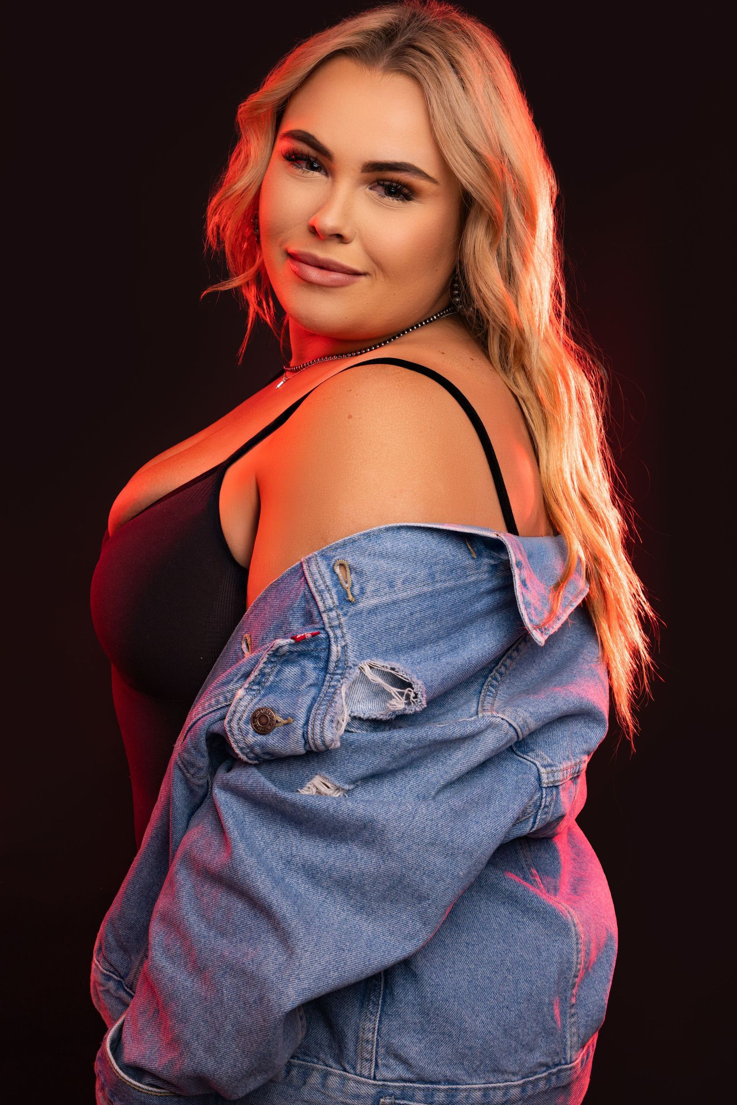 Model in denim jacket poses against dark background with pink lighting effects.