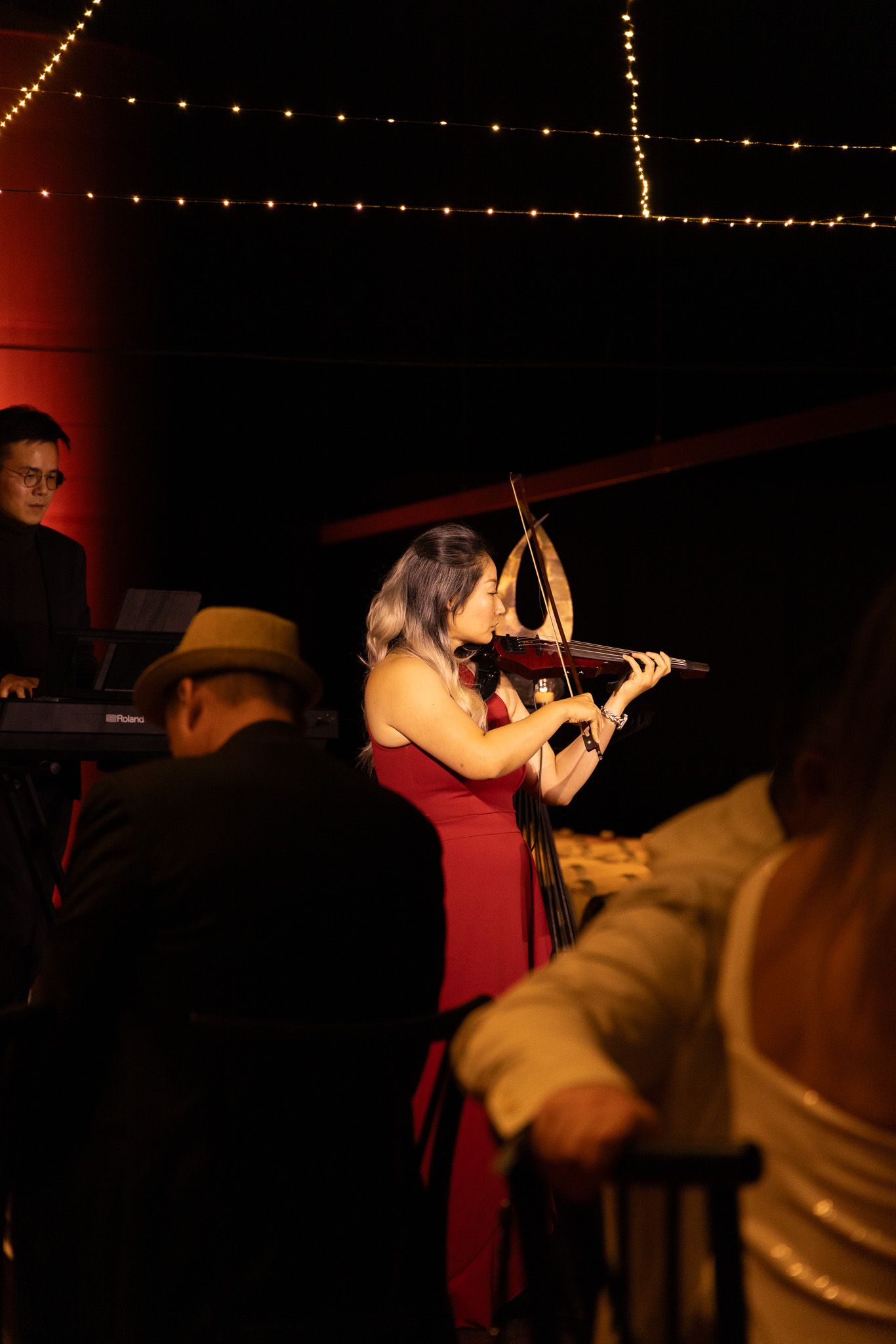 Musicians perform on stage in a dimly lit venue with string lights visible in the background.