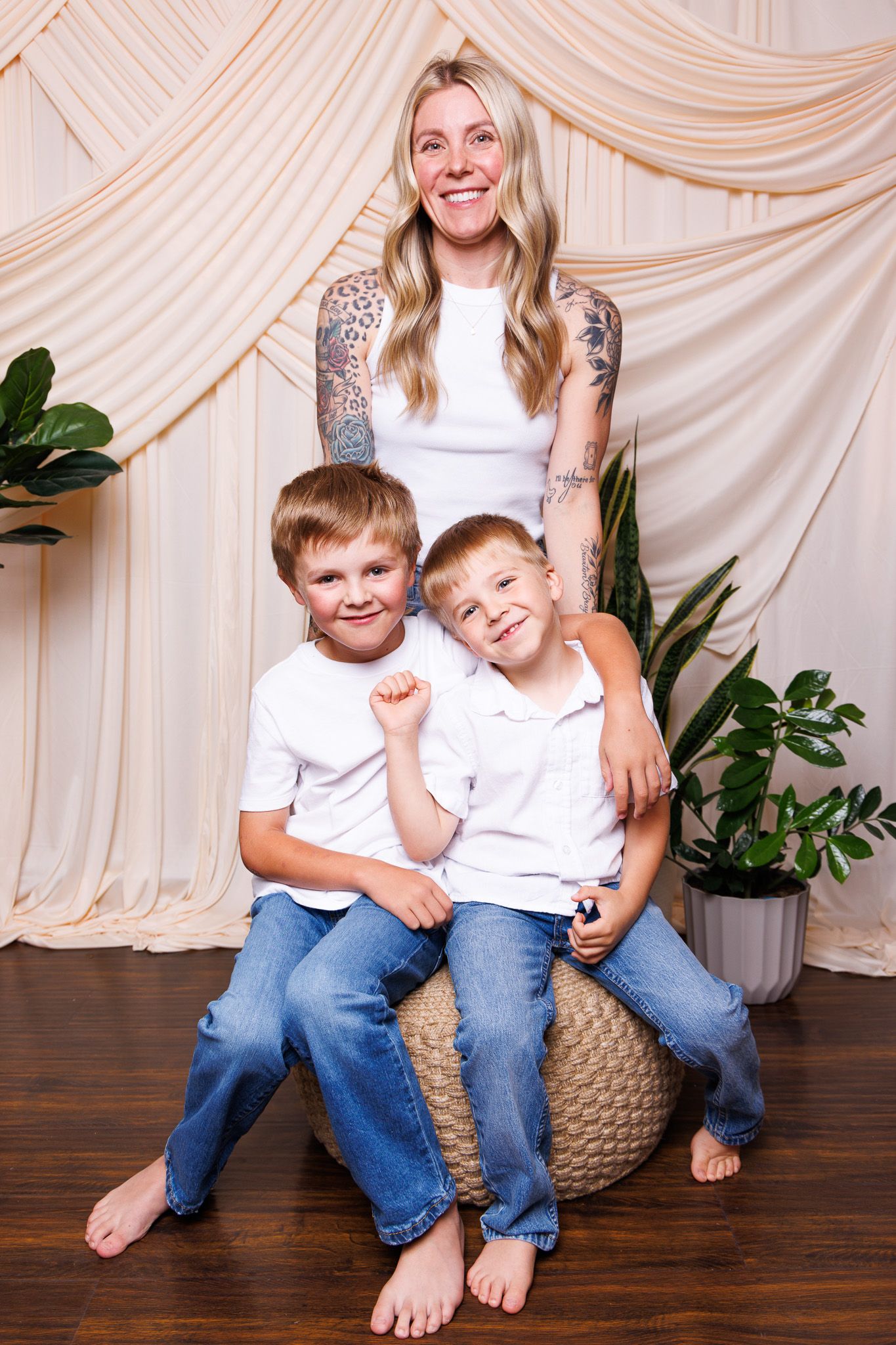 A smiling blonde tattooed mom poses with her two young sons in white shirts and jeans against a cream draped backdrop.