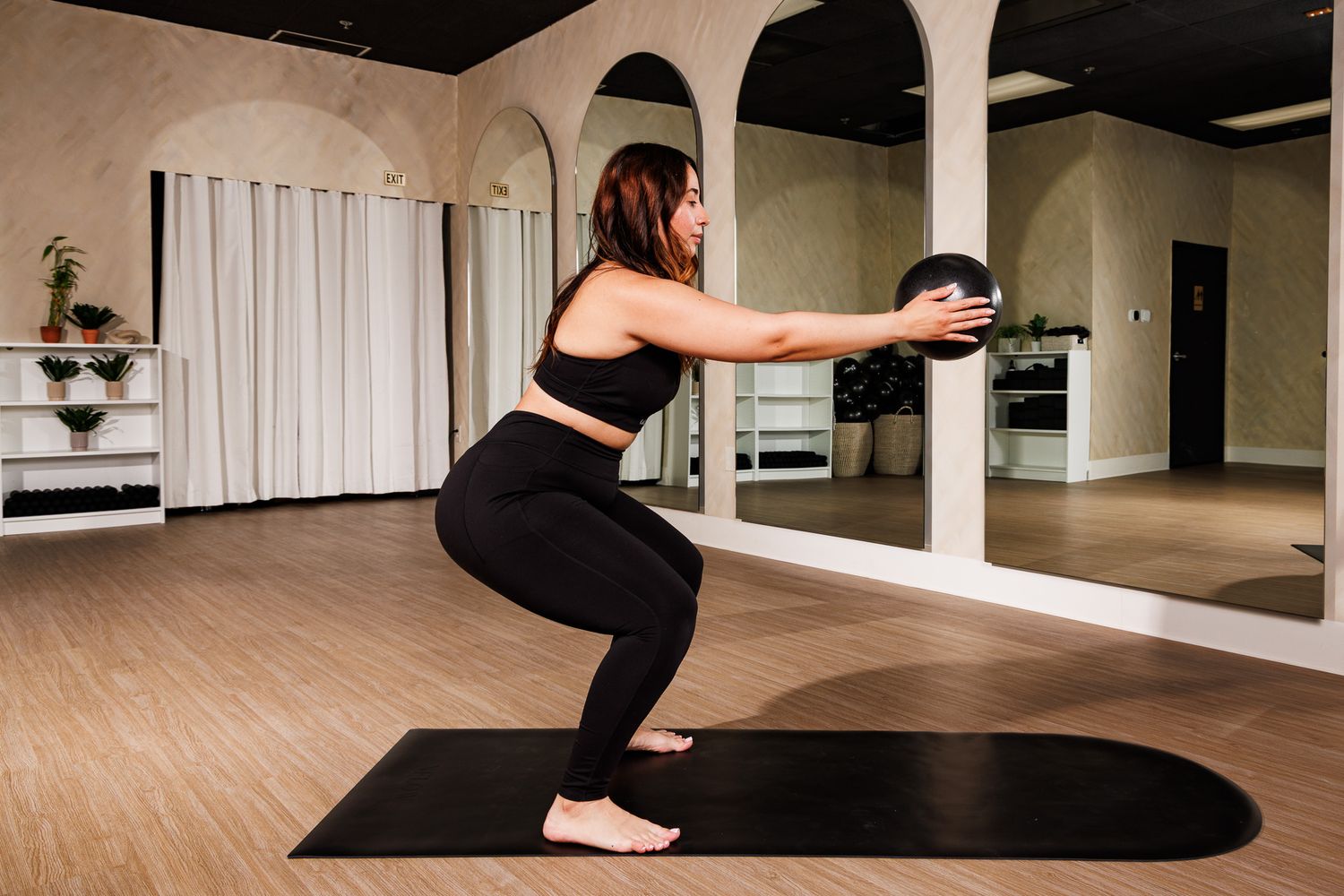 Woman in black activewear performing a squat with medicine ball in a bright mirrored studio.