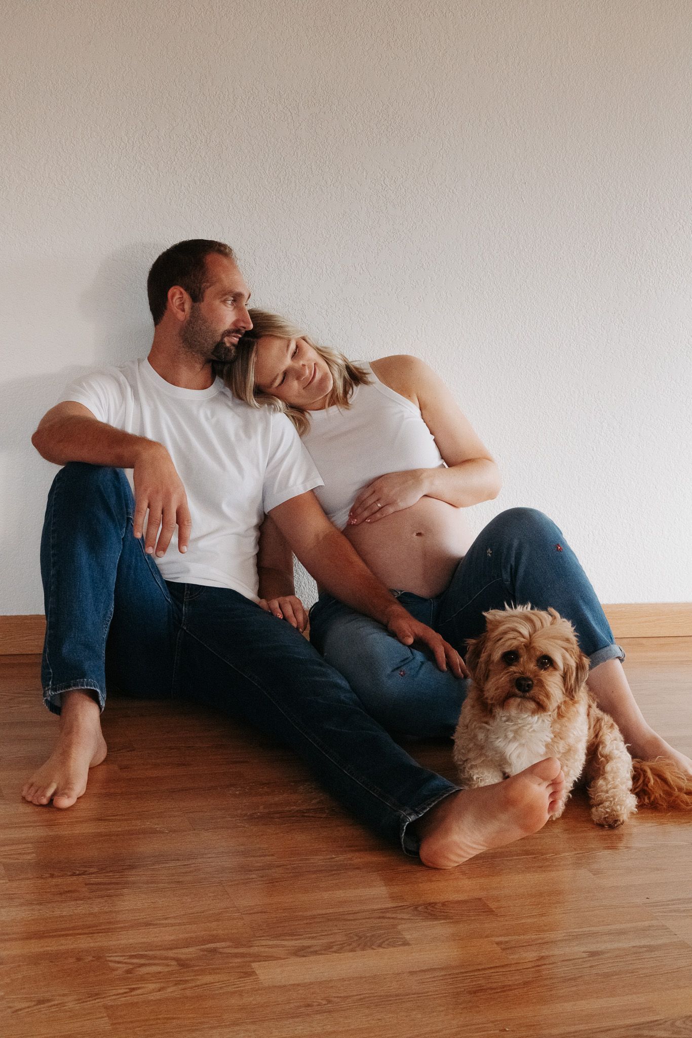 Expecting couple sits on hardwood floor with their small fluffy dog in casual home setting.