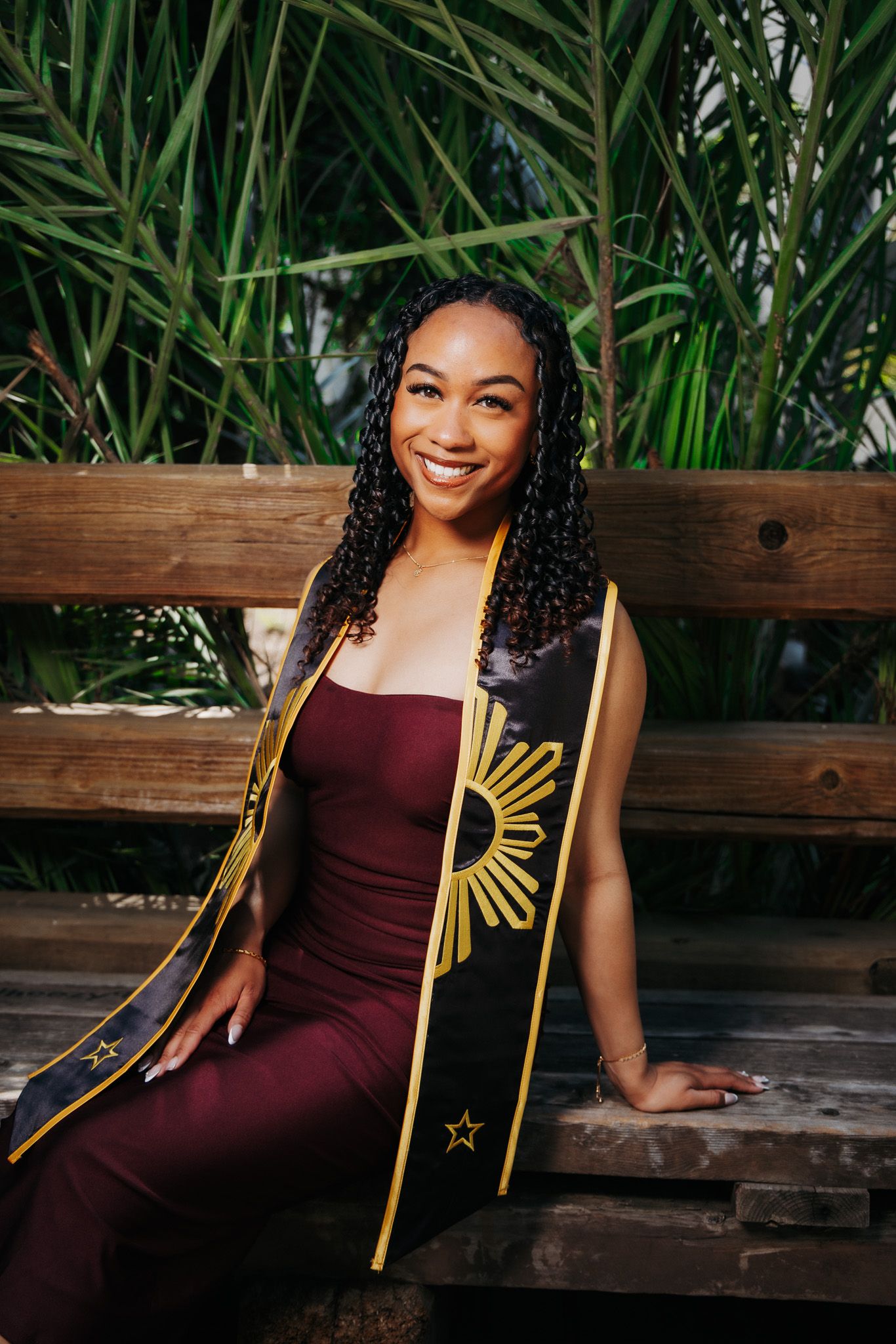 A graduate in a burgundy dress and gold sash sits on a wooden bench surrounded by tropical greenery for graduation photos.
