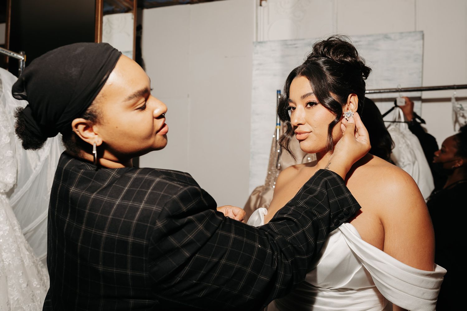 A stylist in a plaid jacket adjusts details on a person wearing a white off-shoulder dress.