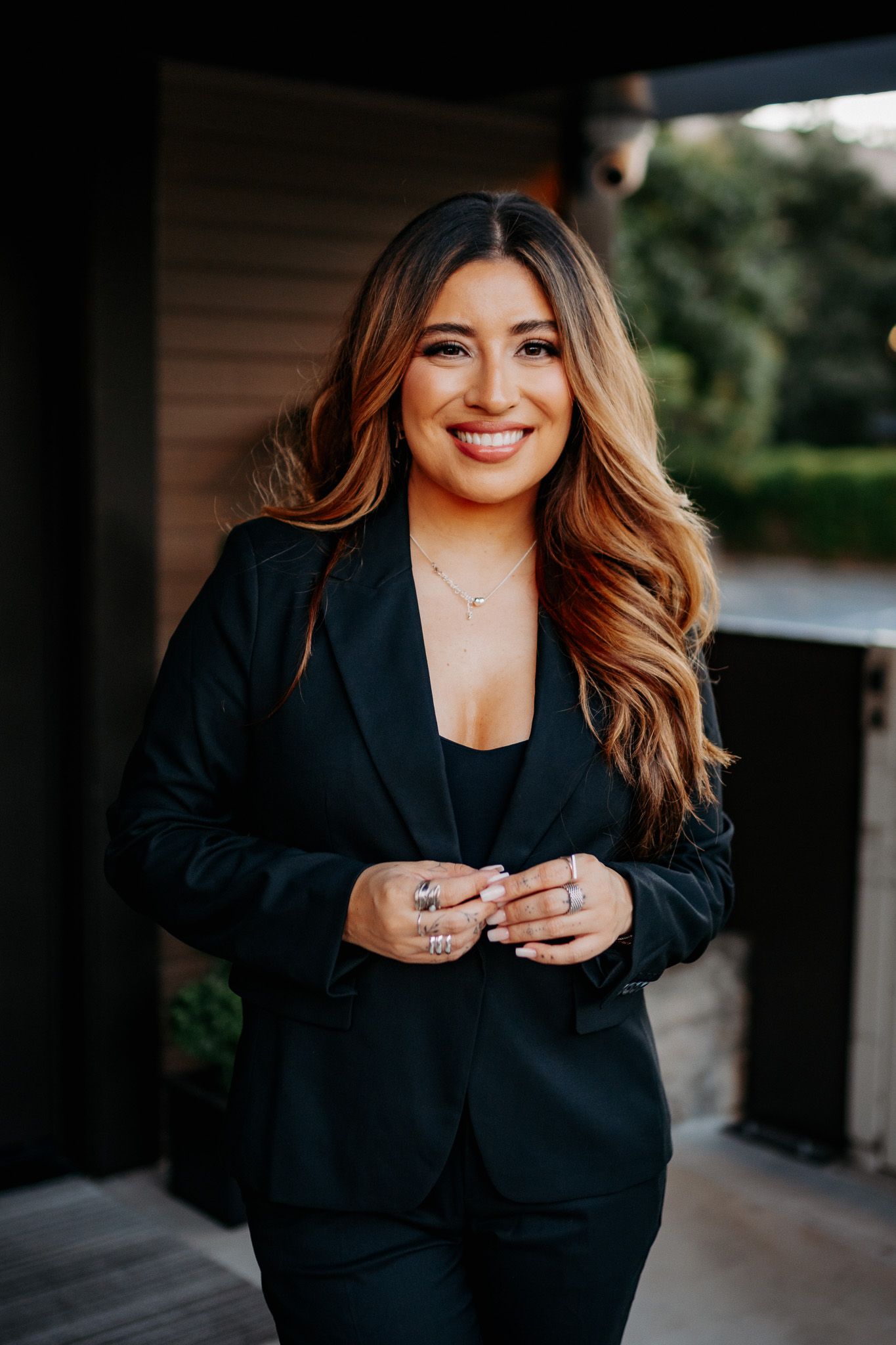 Professional portrait of someone in a black business suit with caramel-colored hair smiling on an outdoor porch.