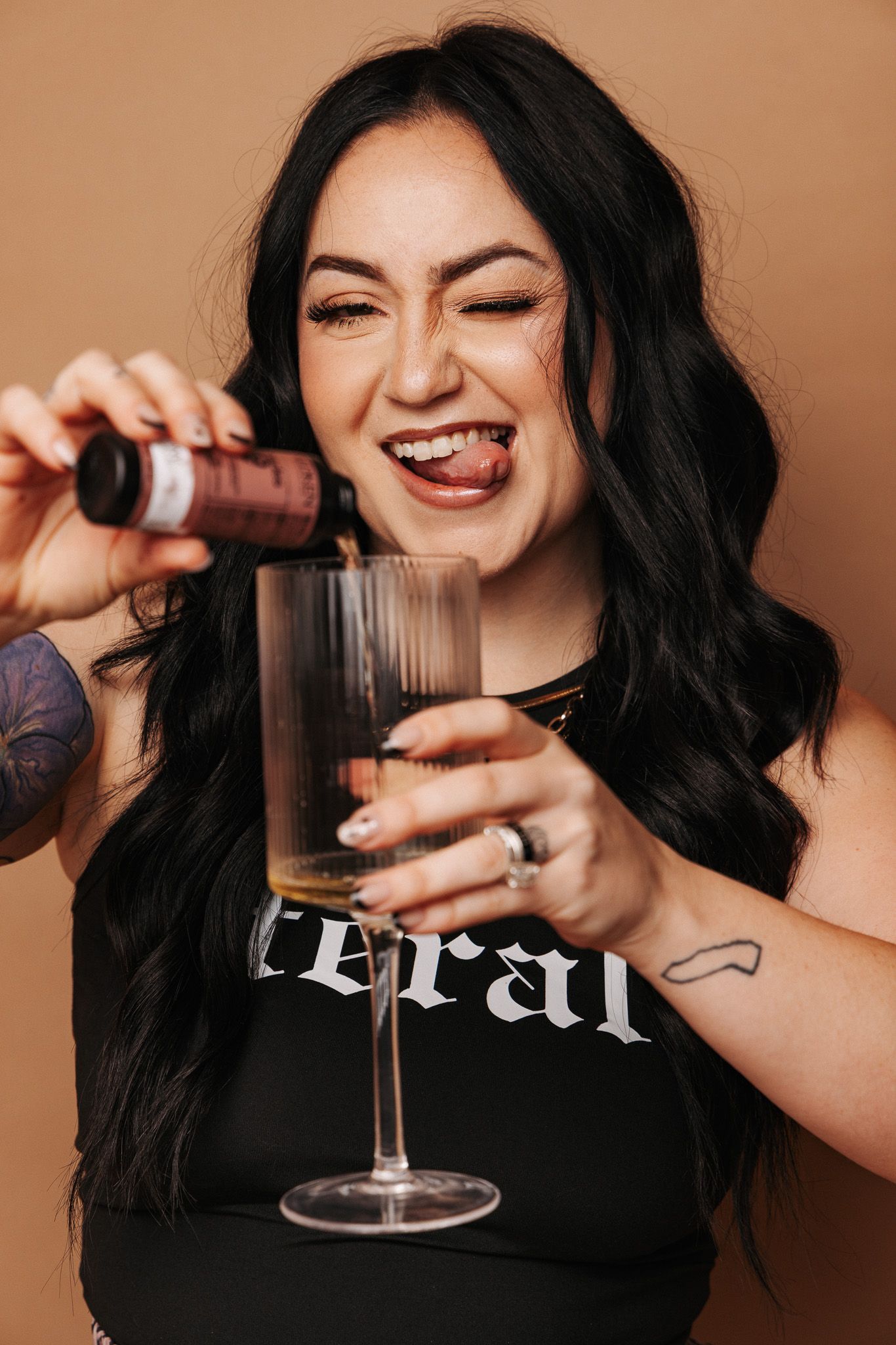 Woman with long dark hair laughs while pouring wine into a glass wearing a black top and rings.
