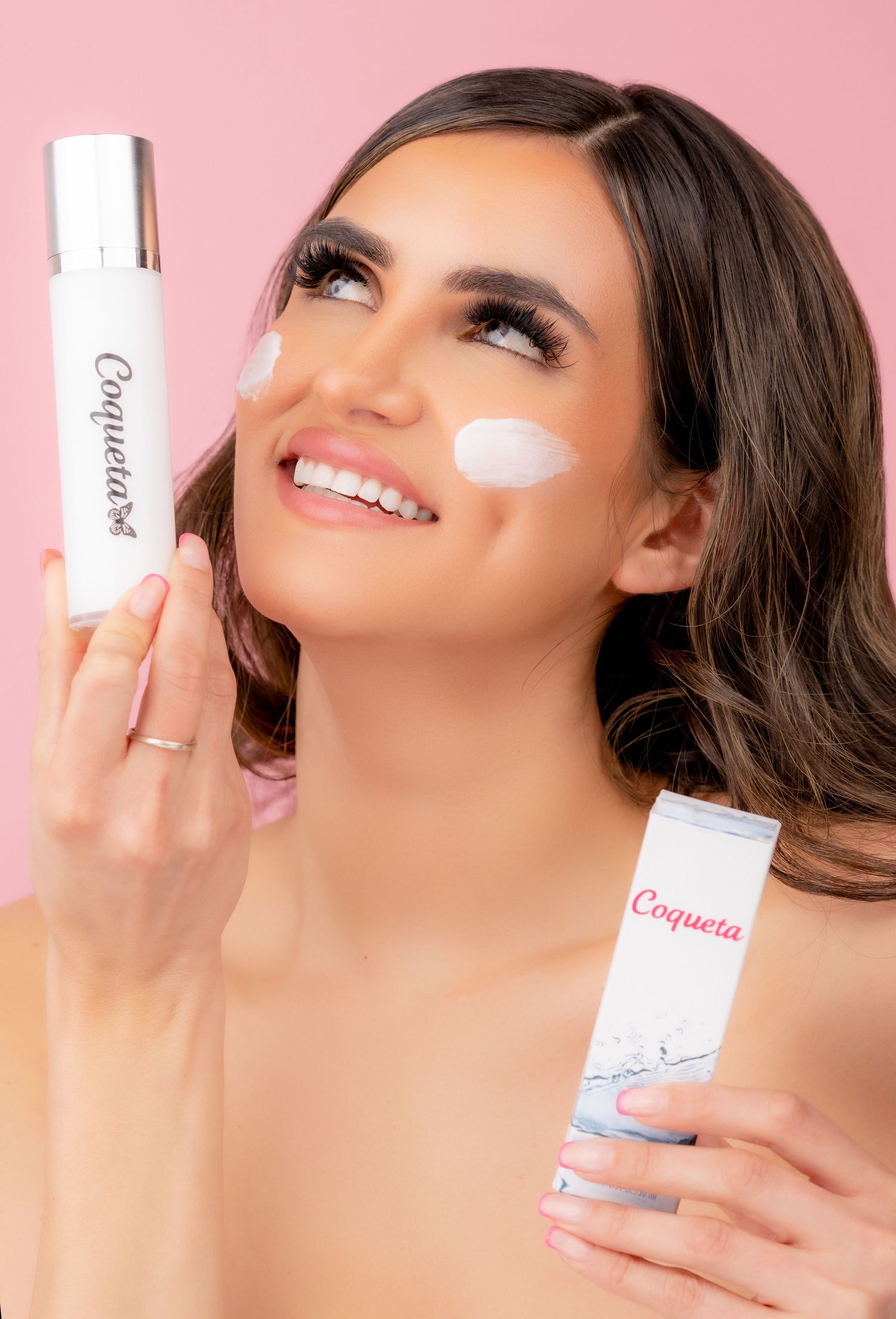 Beauty portrait with skincare products showing moisturizer application on cheeks against pink background.
