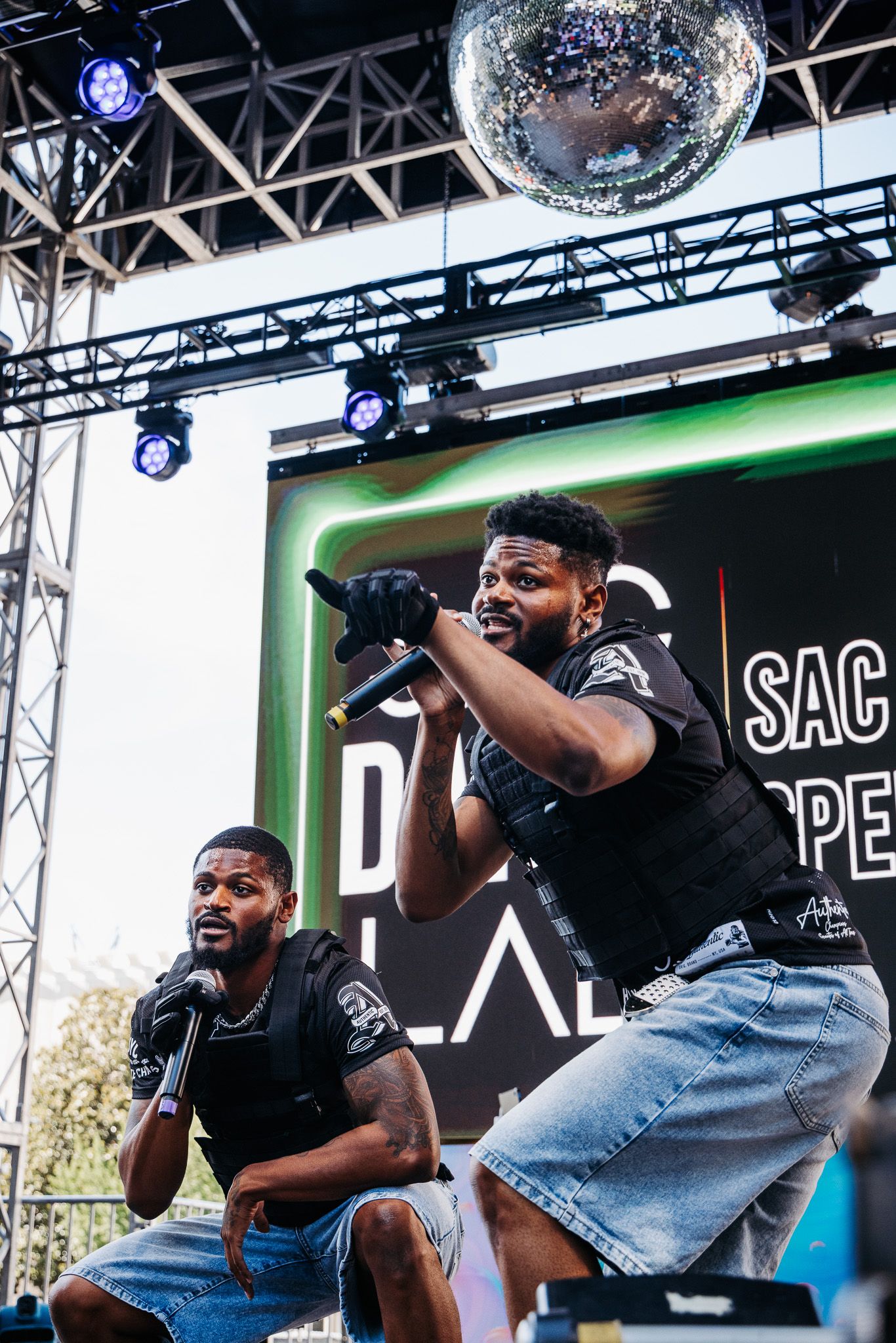 Two performers in black shirts and denim enthusiastically sing on stage under a disco ball and stage lighting.