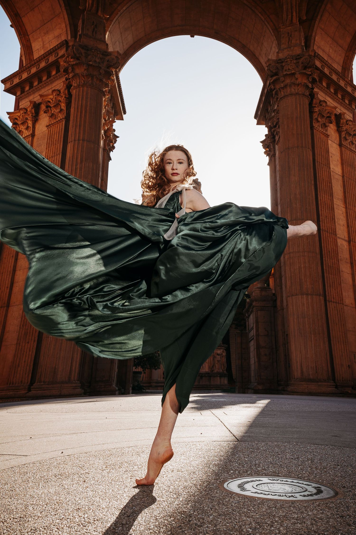 A flowing dark emerald dress dances in the wind against ornate stone architecture at sunset in dramatic fashion.