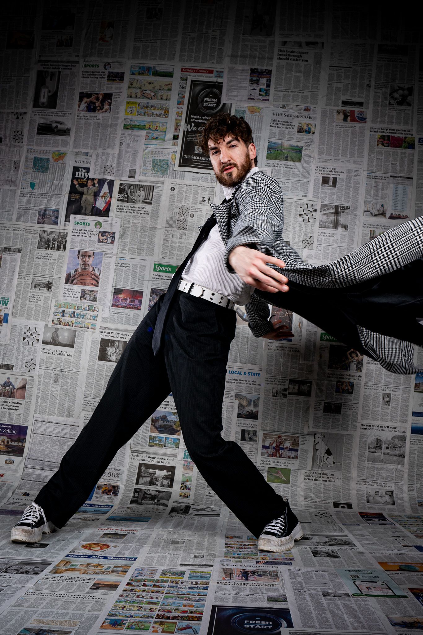 Dynamic pose of someone in business attire doing an energetic dance move against a wall covered in newspaper pages.