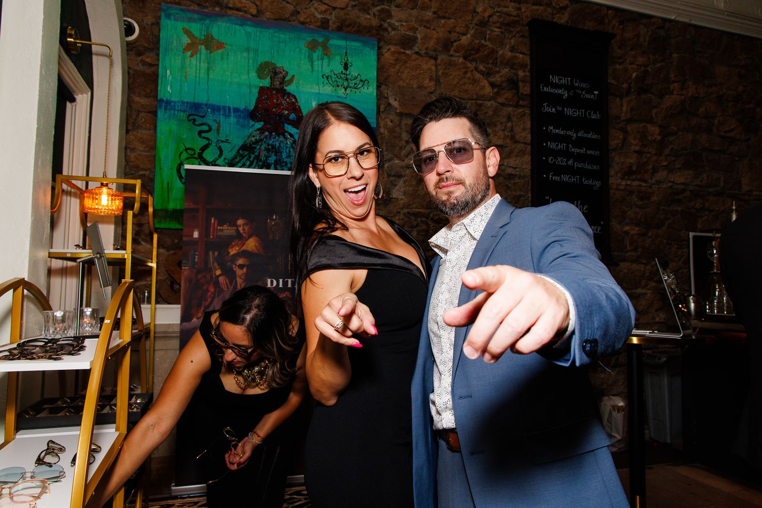 Two people pointing at camera and smiling at an indoor event, man in blue blazer and woman in black.