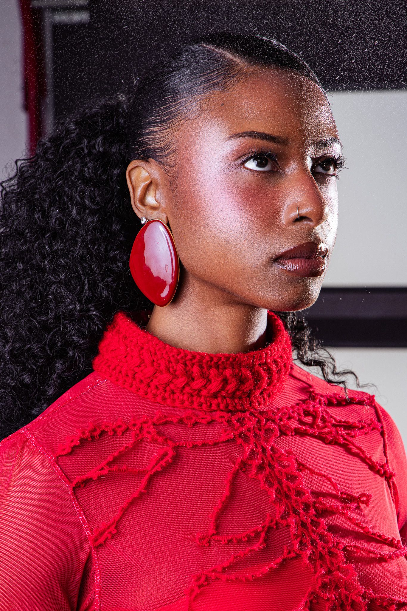 A young Black woman in a red knit turtleneck and large red earrings gazes sideways with a serious expression.