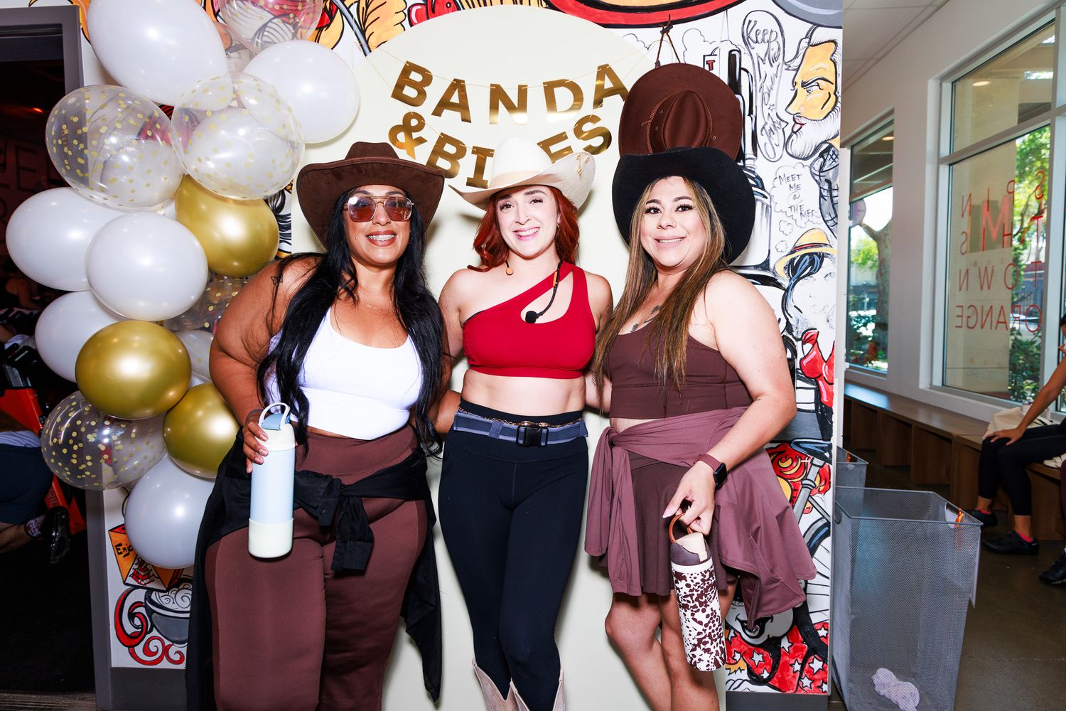 Three women in cowgirl outfits pose together at a Banda and Bites event with balloon decorations.