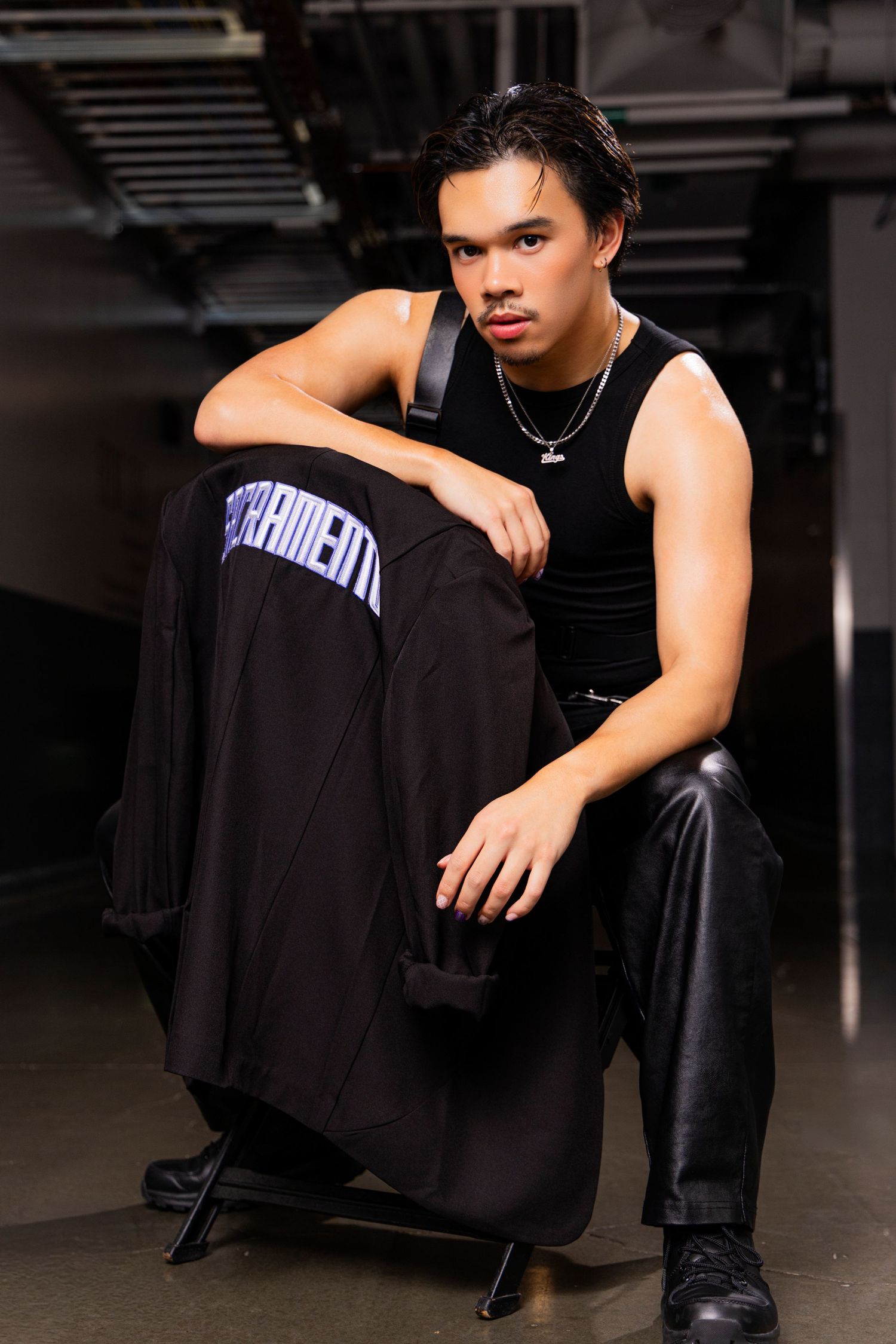Young man in black tank top and leather pants sits confidently in an urban setting wearing chain necklace.