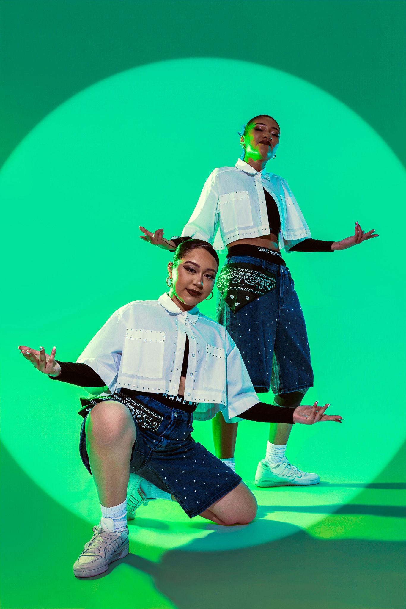 Two models pose dynamically in matching white crop tops and denim skirts against a vibrant green background.