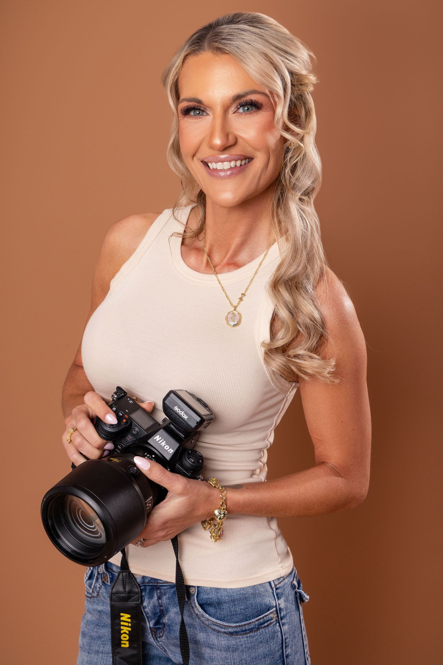 Professional photographer in white tank top and jeans holding DSLR camera against brown studio backdrop.