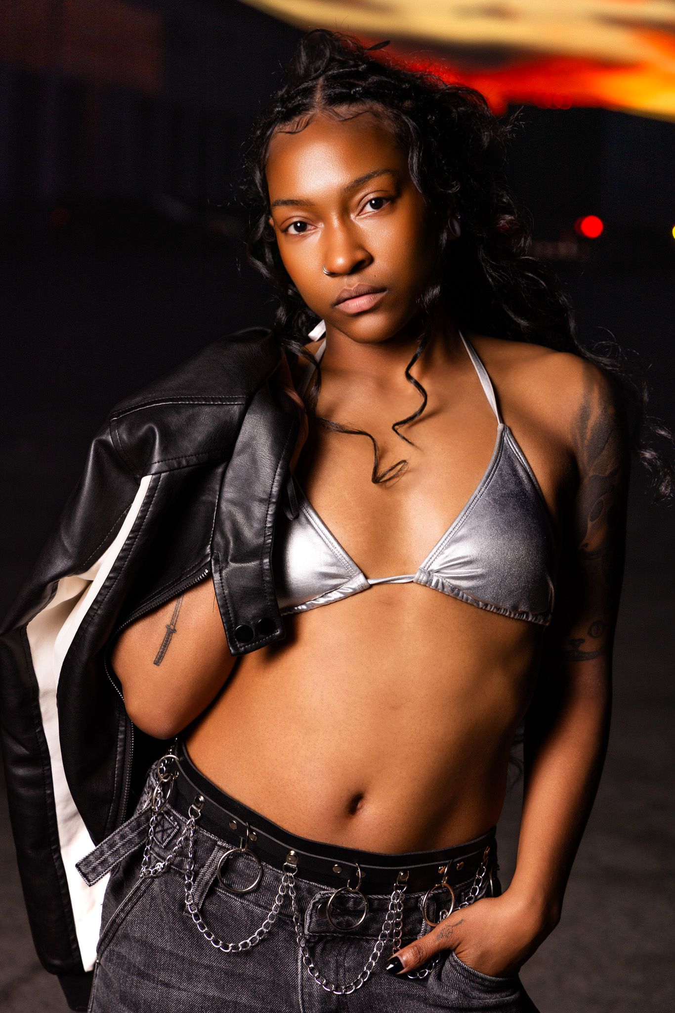 Fashion model in silver bikini top and black leather jacket poses against dramatic sunset lighting.