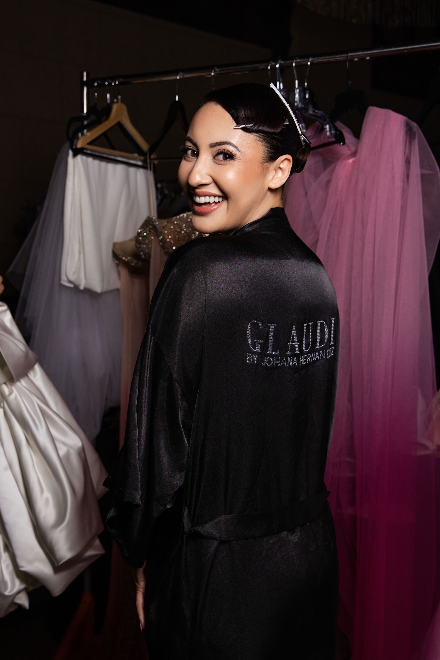 A person wearing a black Glaudi robe smiles while standing in a dressing room with wedding dresses visible in background.