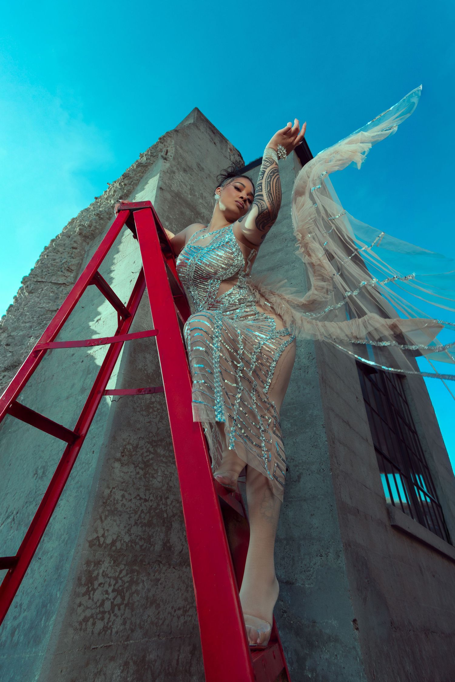 A figure in a flowing dress poses against a concrete wall with red architectural elements under a bright blue sky.