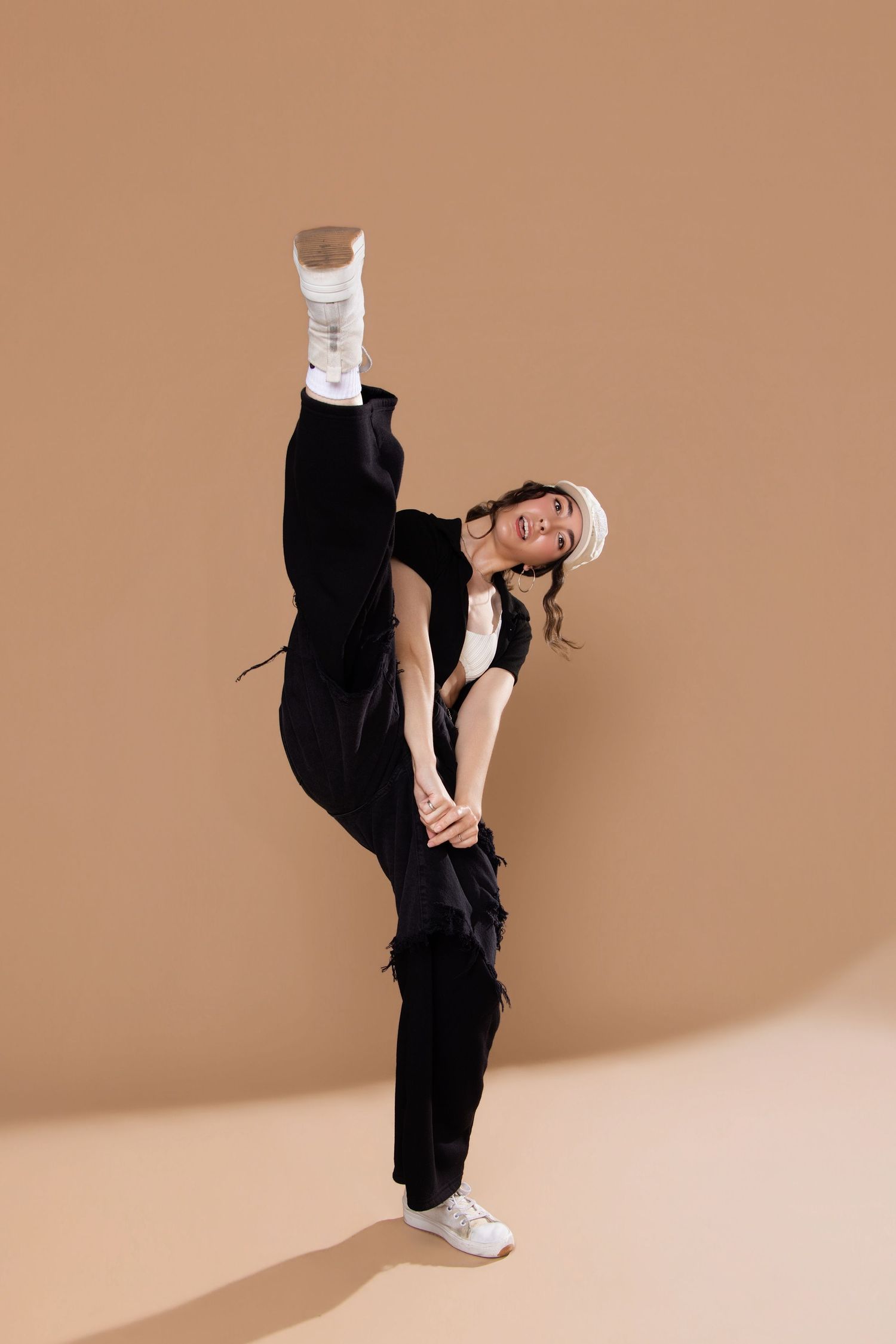 Young woman in black outfit and white hat performs high kick dance move against beige background.