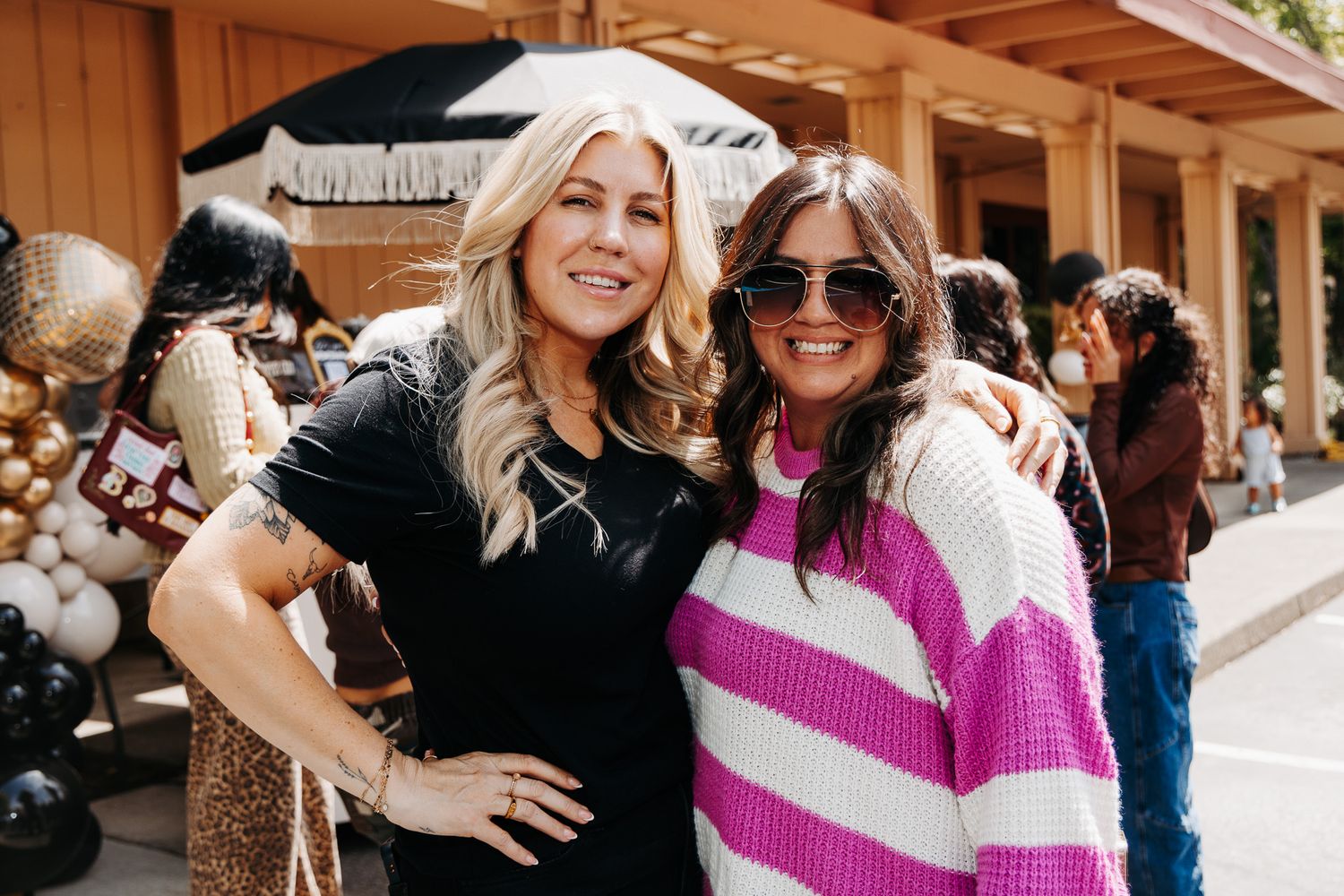Two women smiling and posing together outdoors at a casual event, one in black and one in a pink striped sweater.